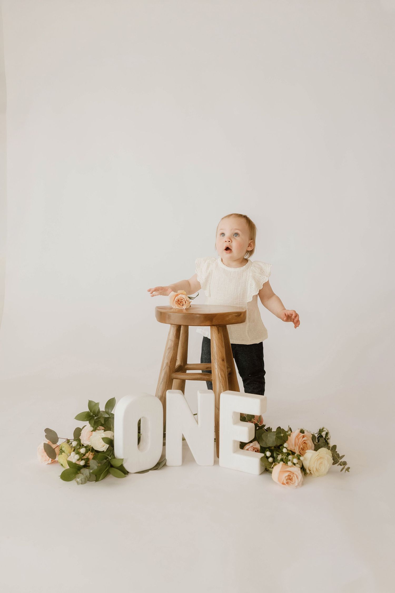 A baby in white clothing sits beside large white 'ONE' letters and floral decorations in a minimalist photo series.