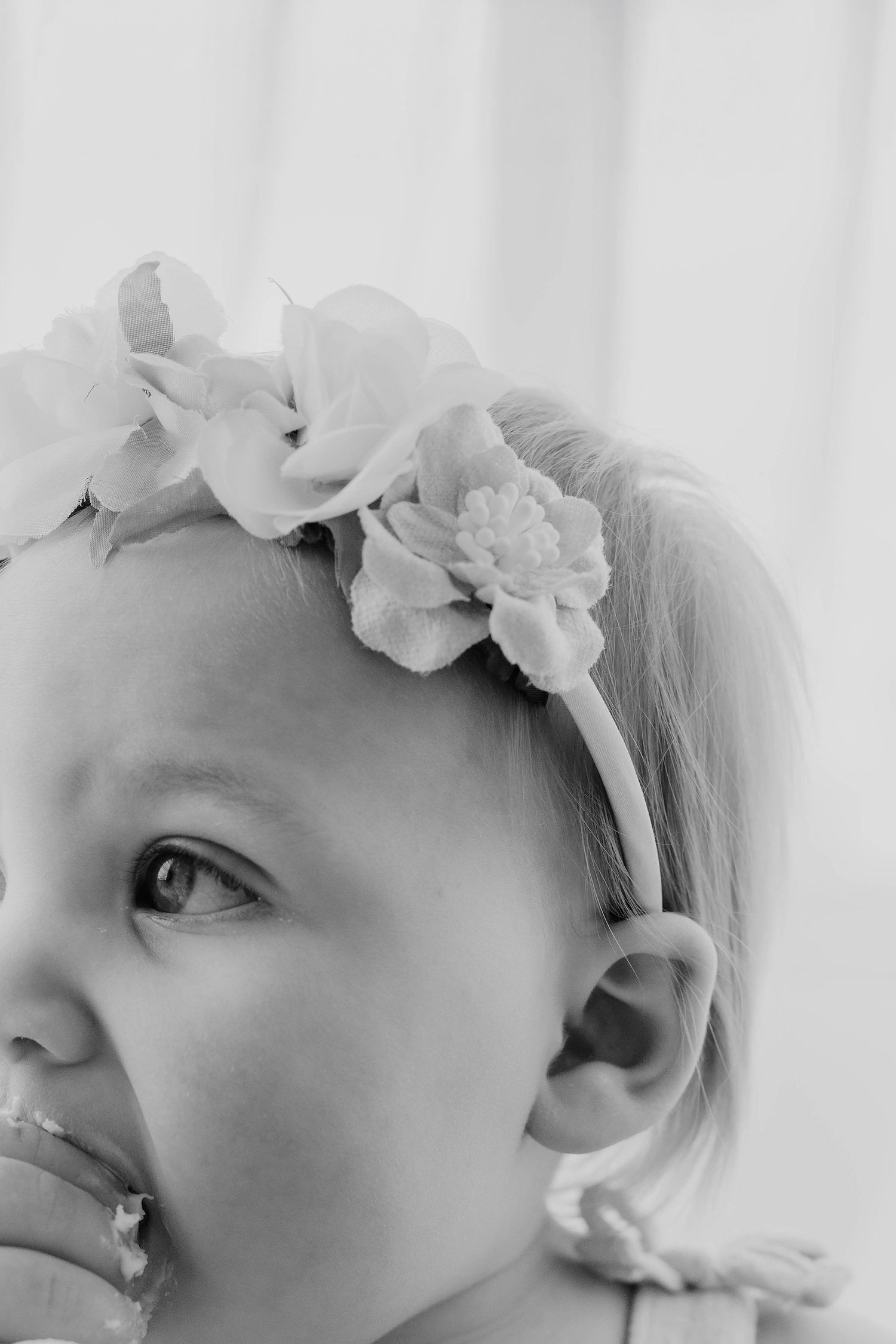 Close-up black and white portrait of a baby girl wearing a delicate floral headband with cake on her face.