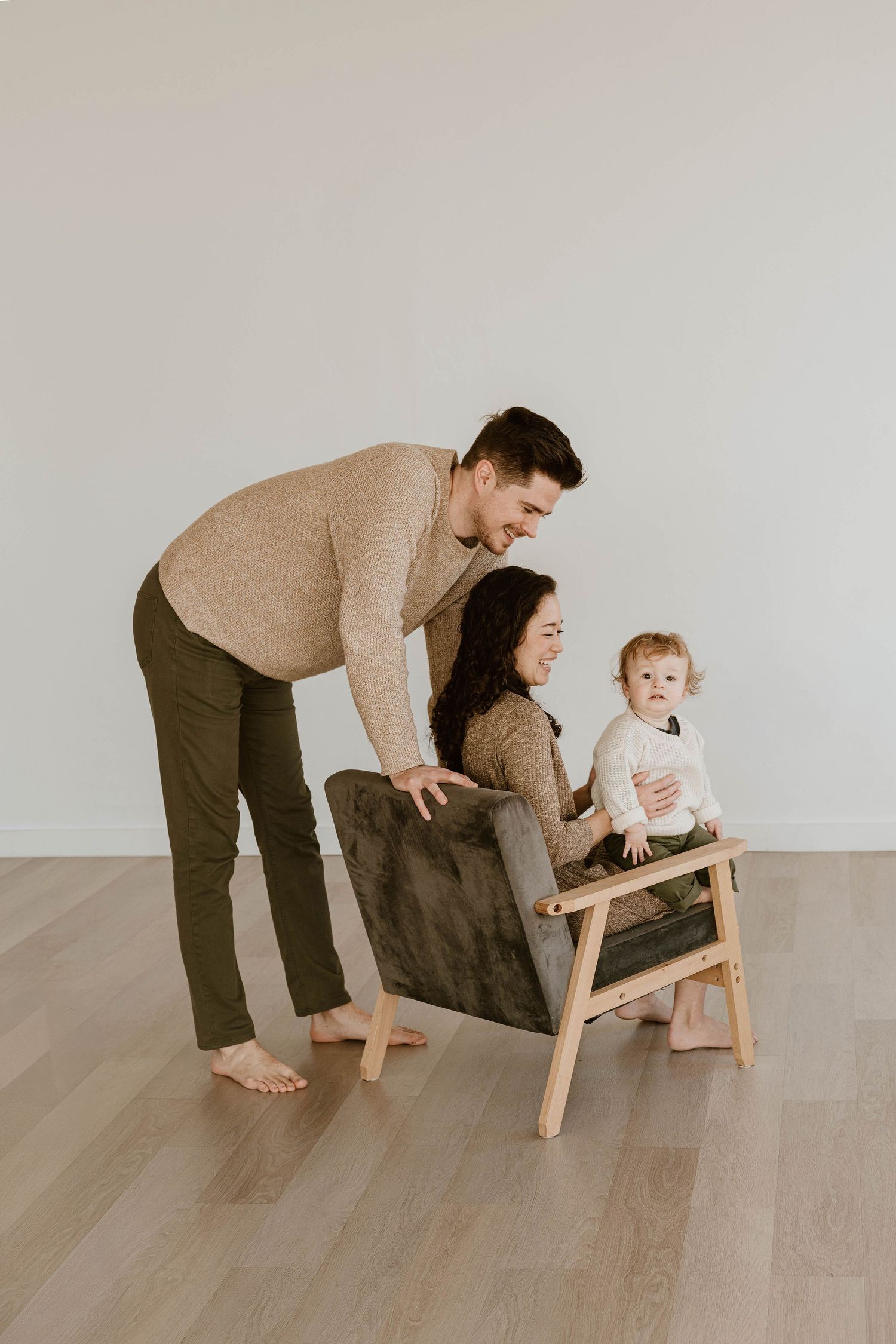 Family playing together on wooden floor with small child climbing on furniture in minimalist setting.