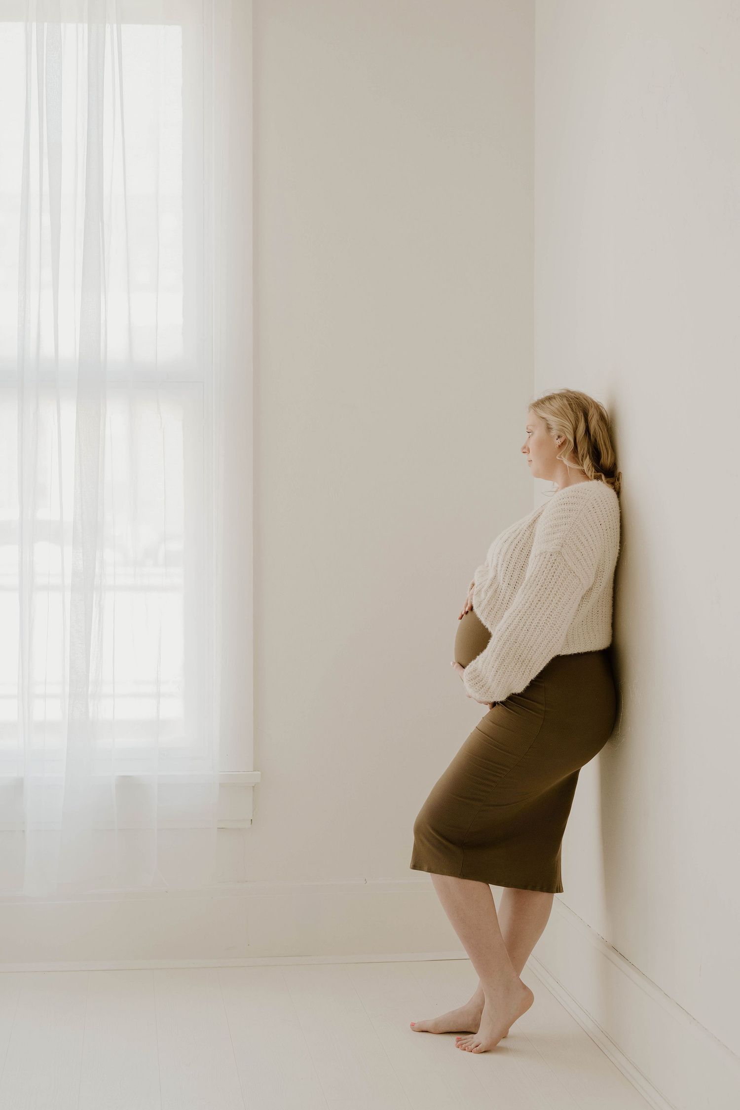 A person in a cream sweater and brown skirt leans against a white wall in a minimalist setting.
