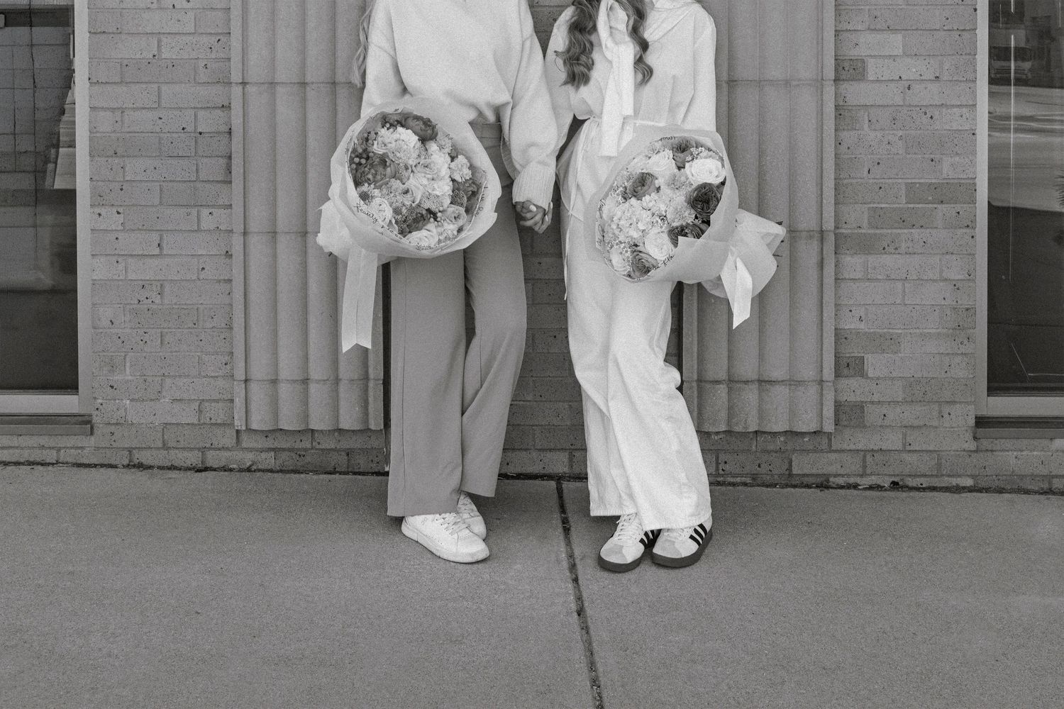 Two brides holding floral bouquets in elegant white outfits pose together outside a brick building in black and white photo.