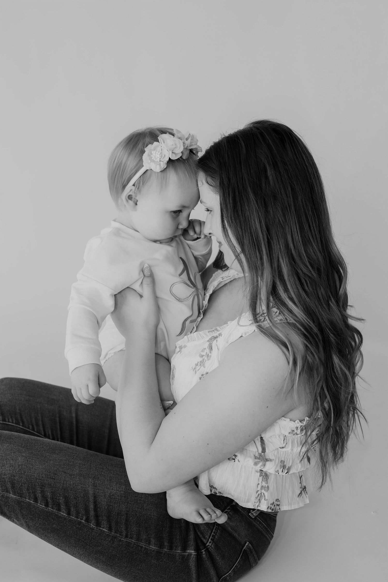 Black and white photo of a mother sitting on the floor, lovingly holding her baby girl with a floral headband.