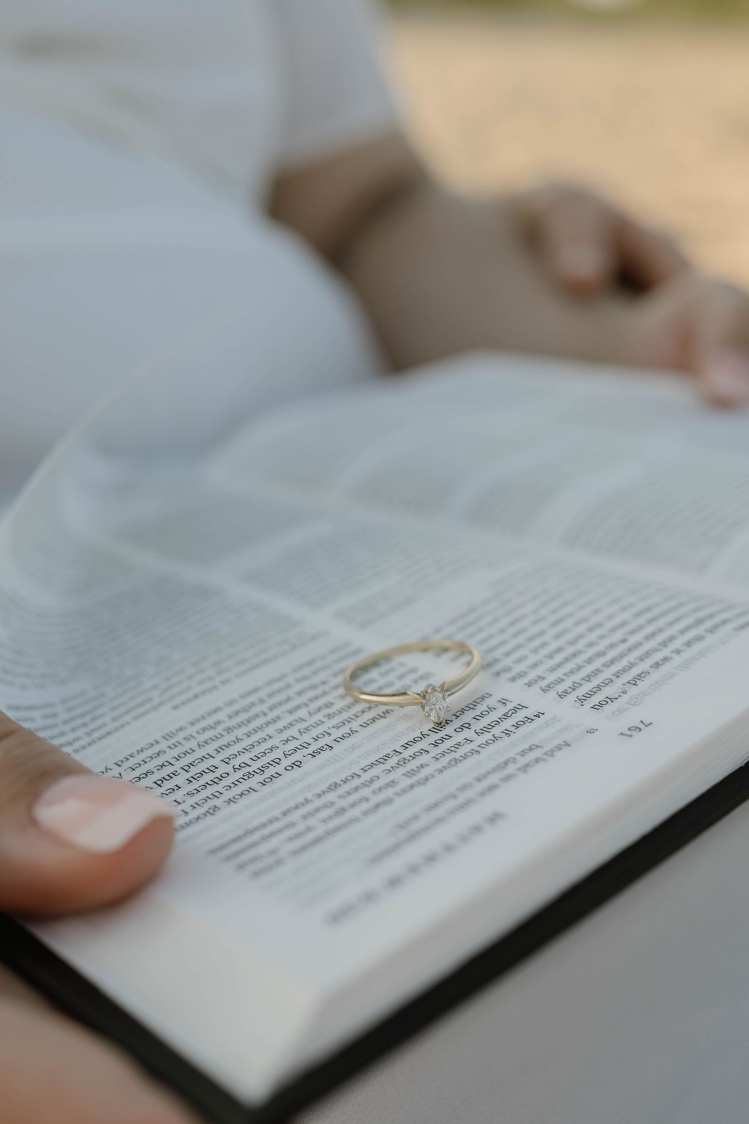 A diamond engagement ring resting on an open book held by a person in a white outfit.