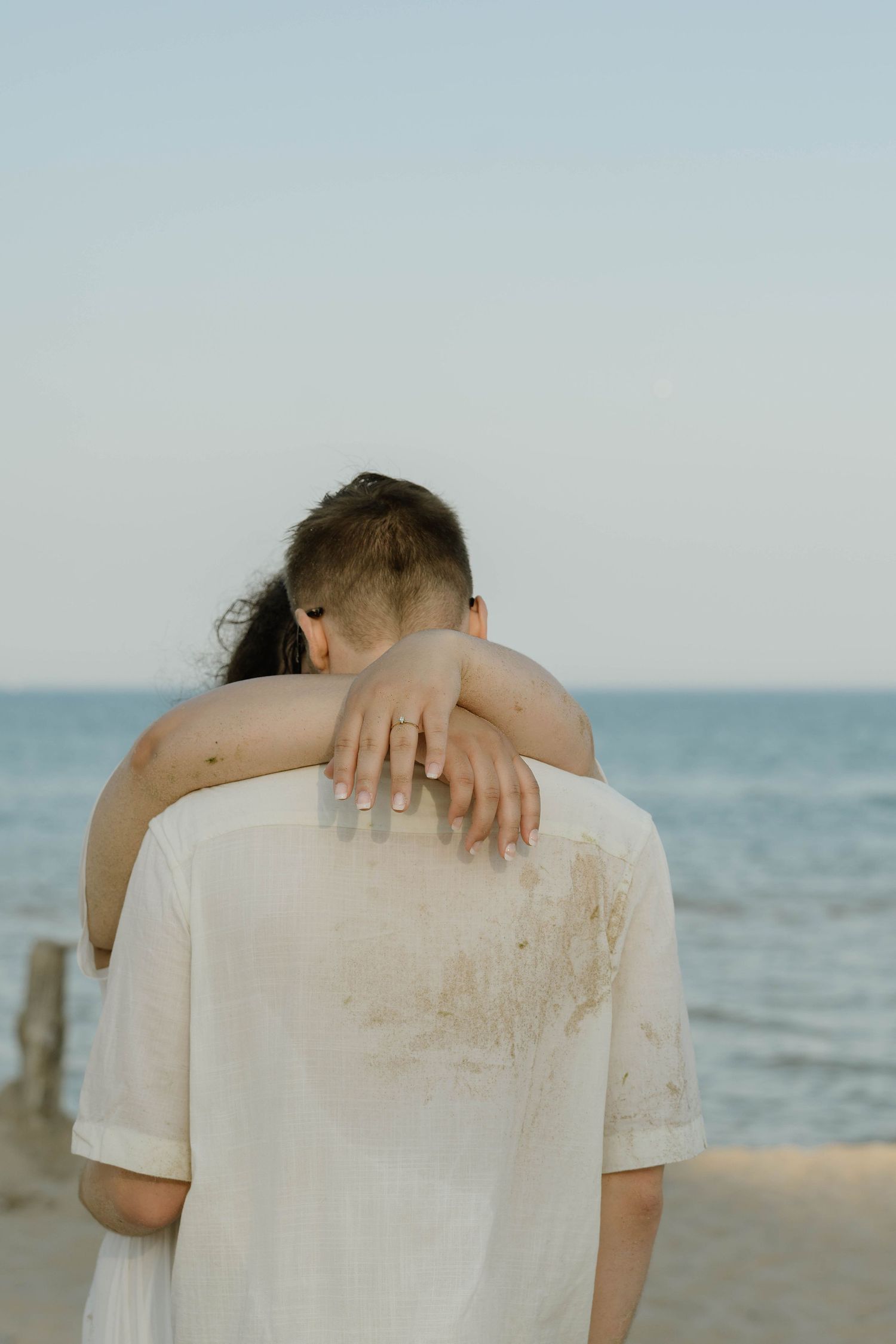 Couple embracing on a beach, woman resting her head on man's shoulder, wearing a ring, with ocean in the background.