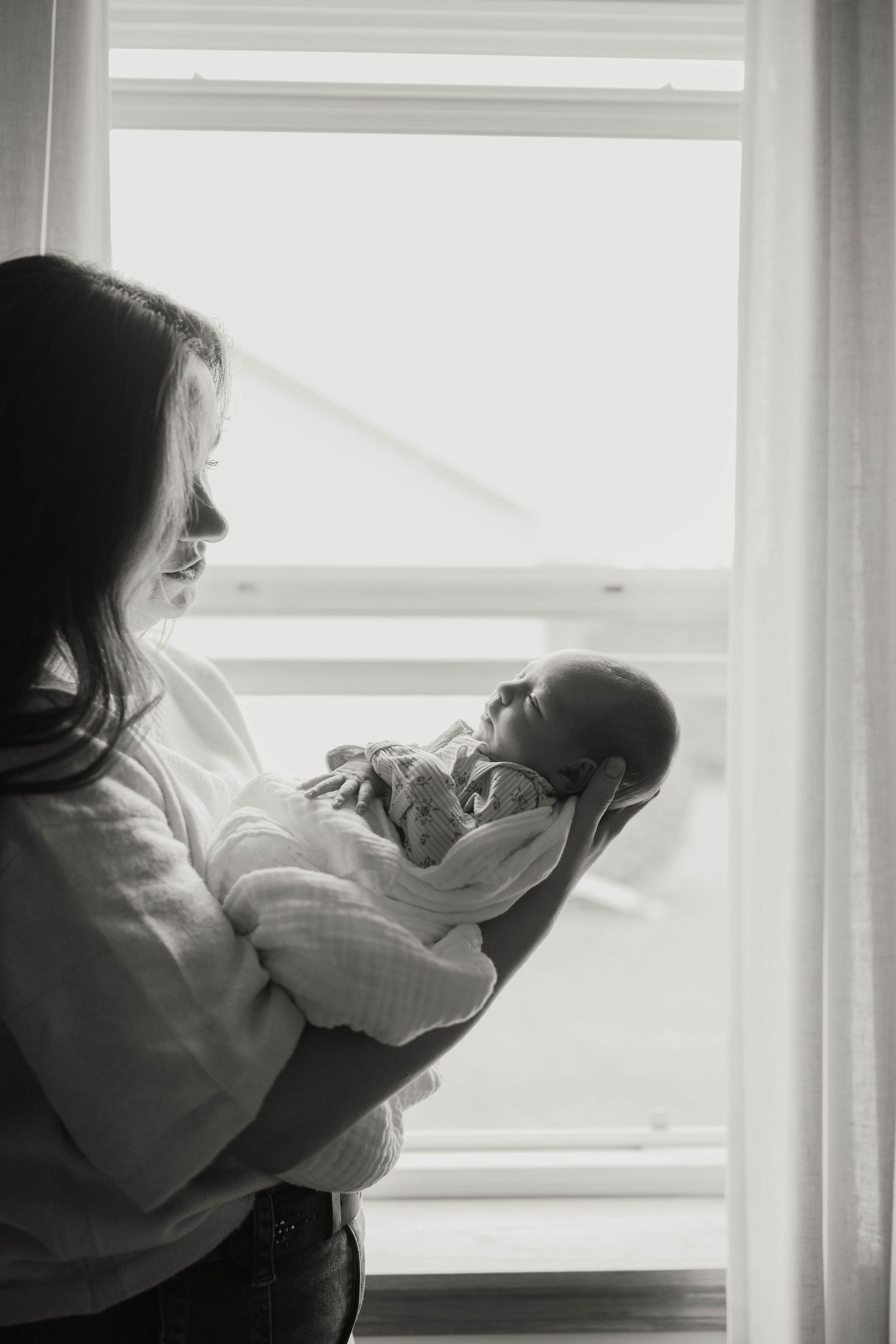 Black and white photo of mother holding newborn baby by a bright window.