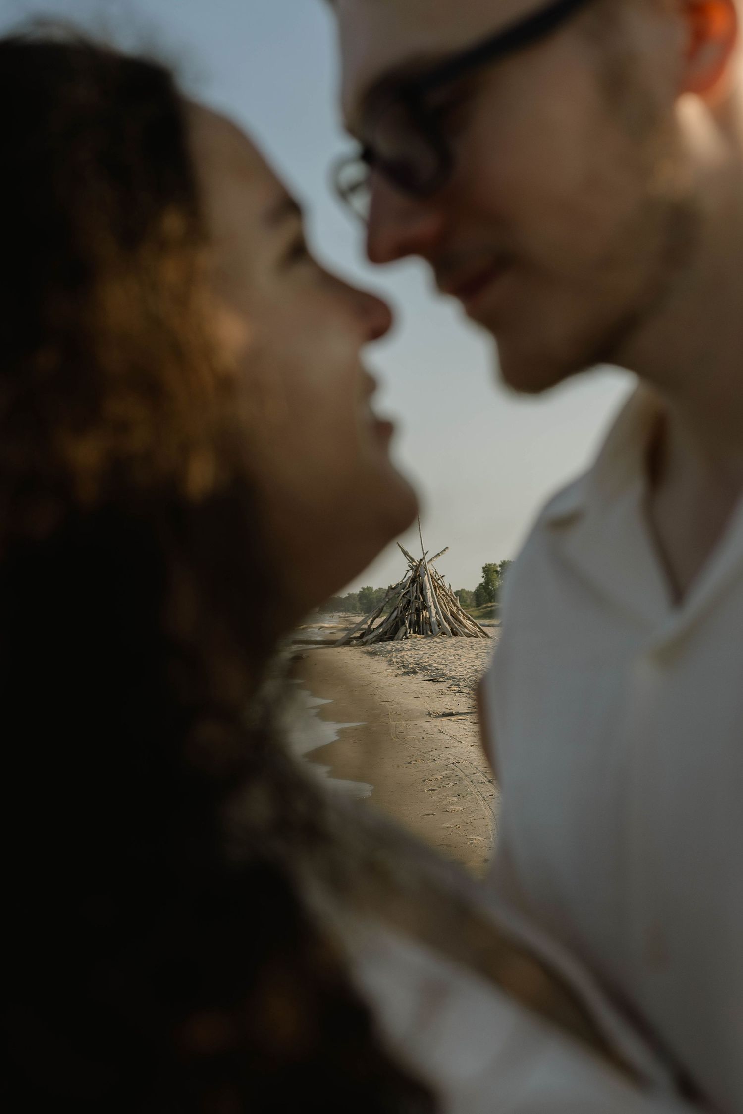 Romantic couple almost kissing on a beach at sunset, with a driftwood teepee structure visible in the background.