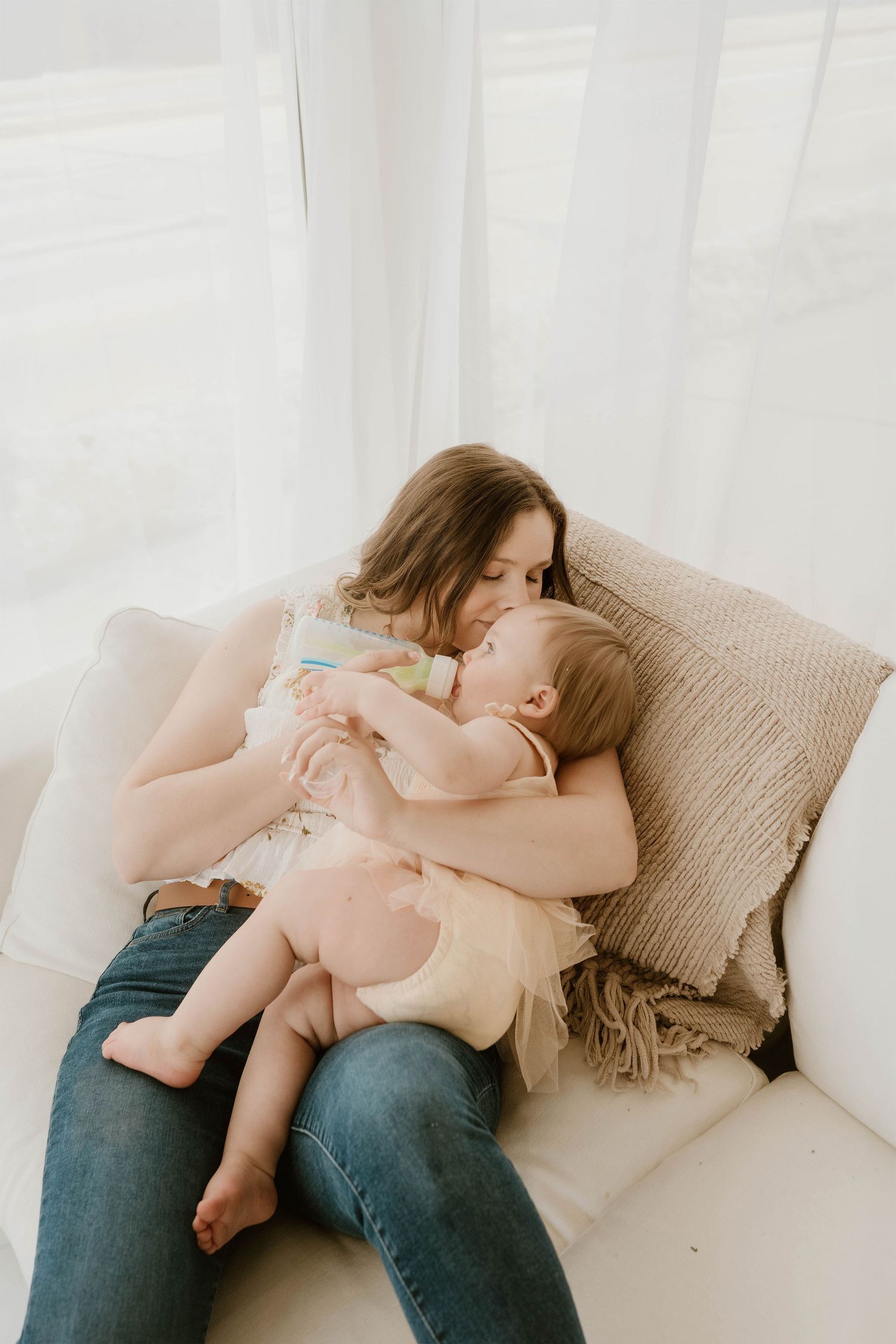 A mother tenderly kisses her baby while bottle feeding on a white couch with neutral pillows.