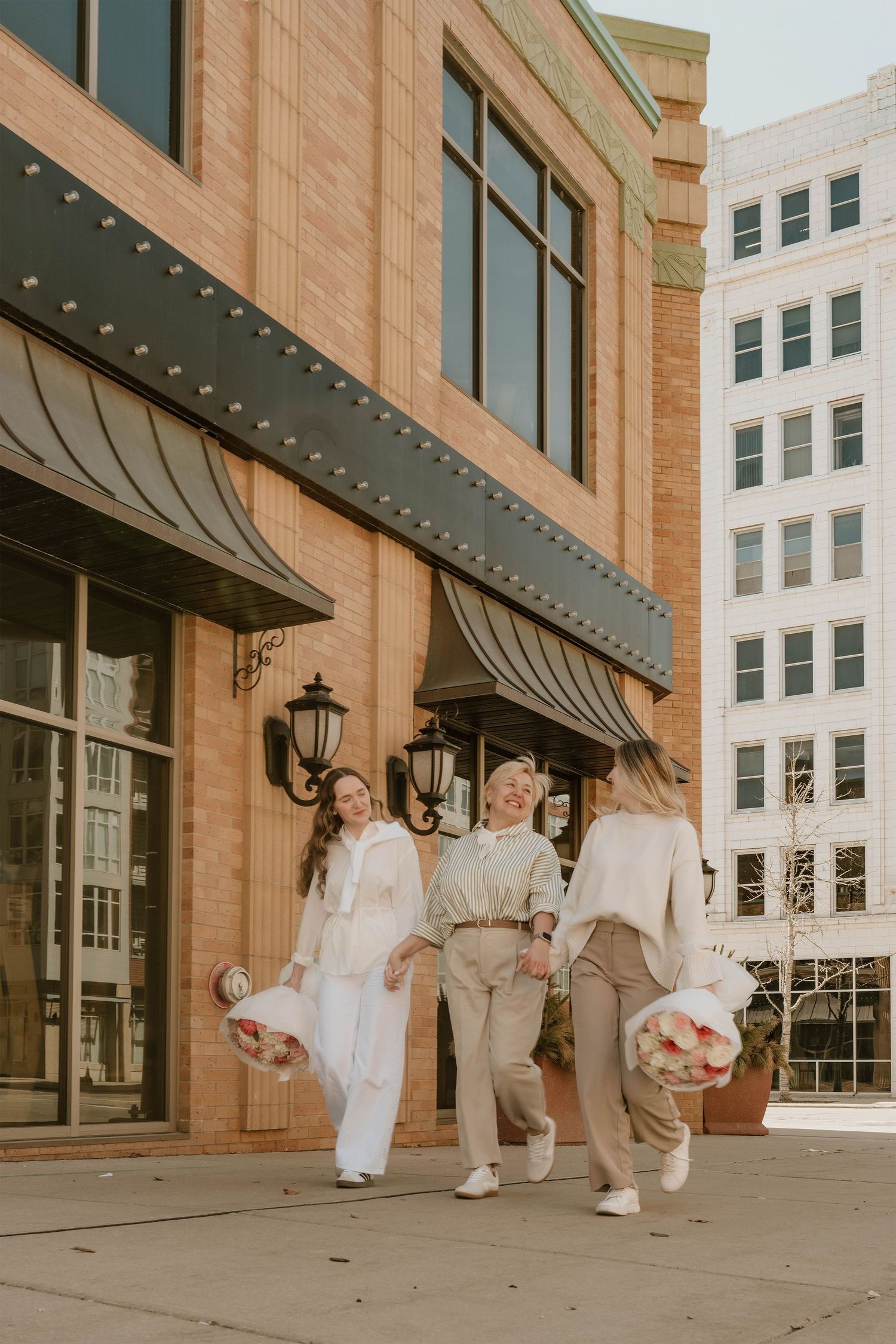Three women in neutral tones walk together outside a brick building, carrying bouquets in an urban setting.
