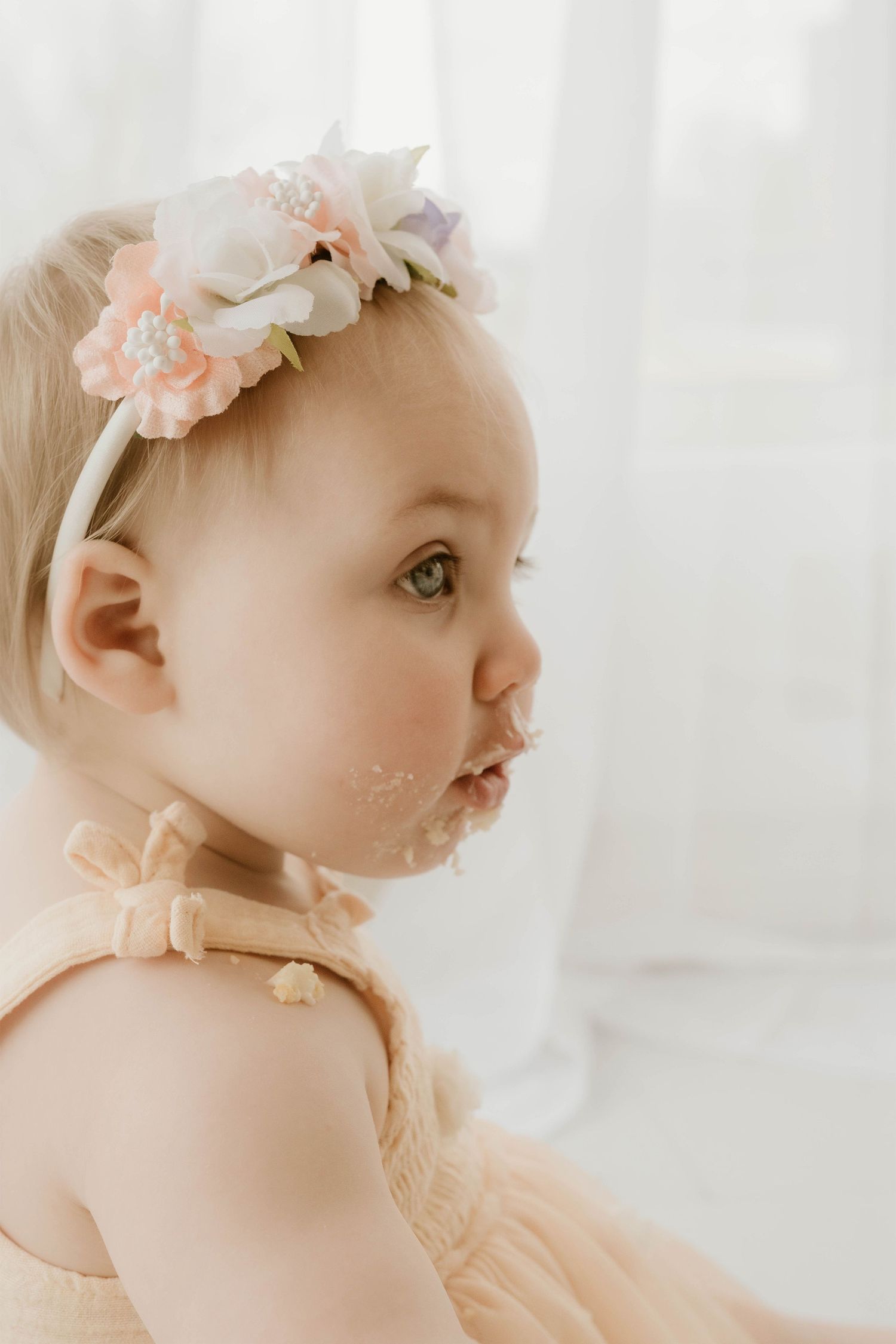 Adorable baby girl in a peach tutu dress and floral headband with cake on her face during first birthday smash.