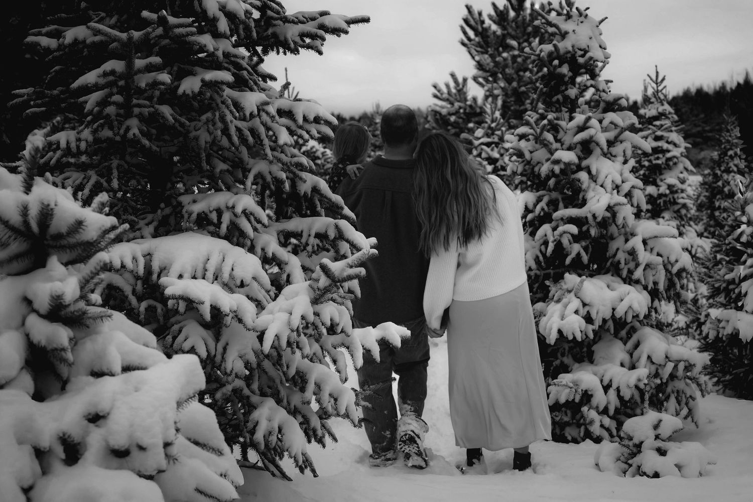 A figure stands in a snowy winter landscape surrounded by snow-covered evergreen trees in black and white.