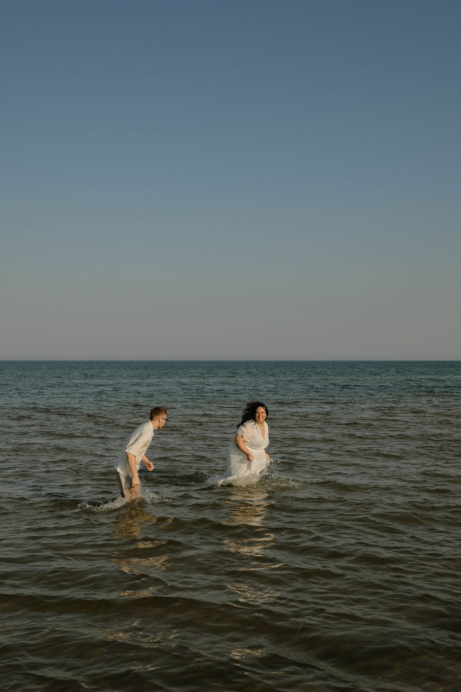 Couple running through shallow ocean water in white outfits under a clear blue sky.