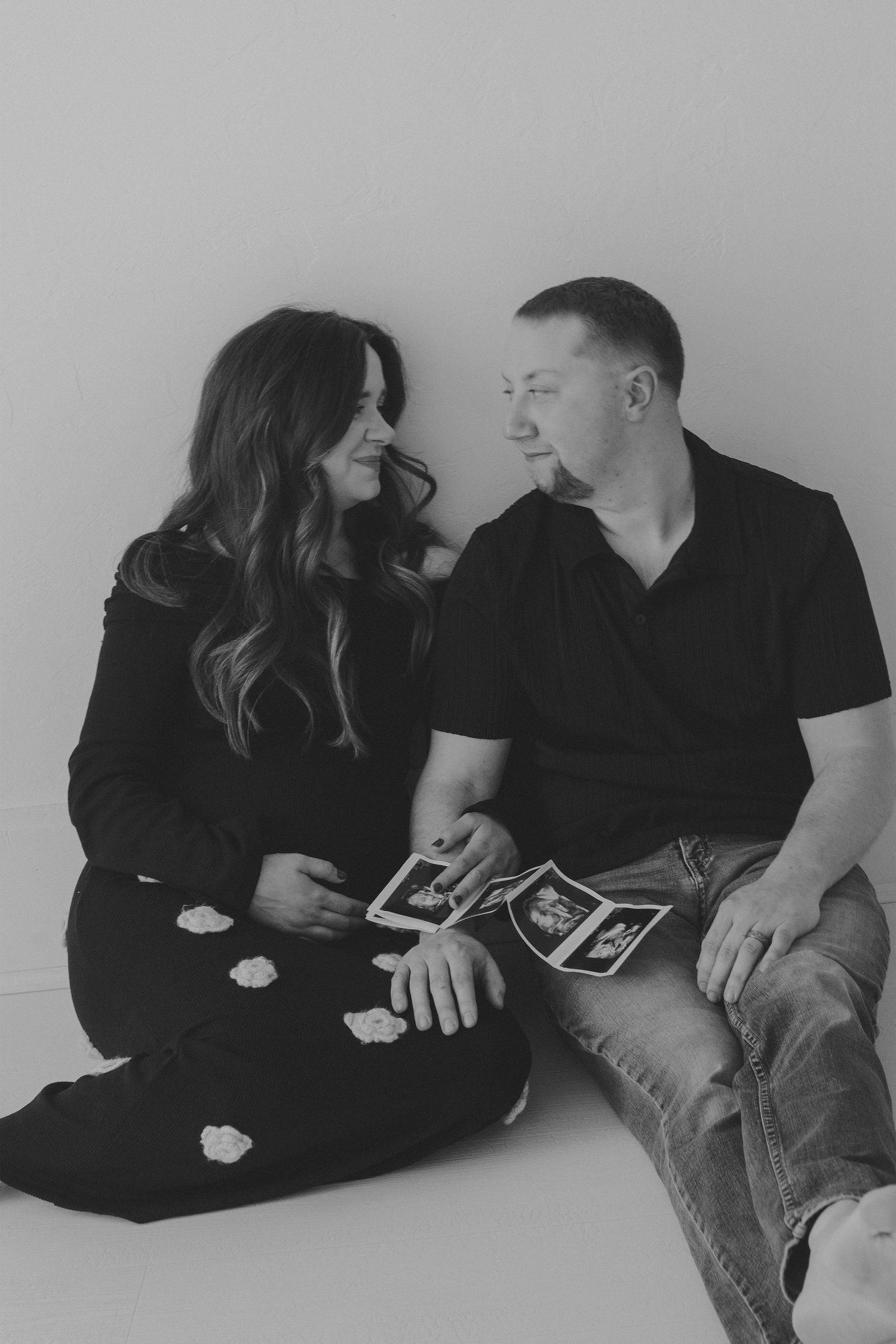 Black and white photo of a couple sitting together holding ultrasound images, smiling at each other during maternity announcement.