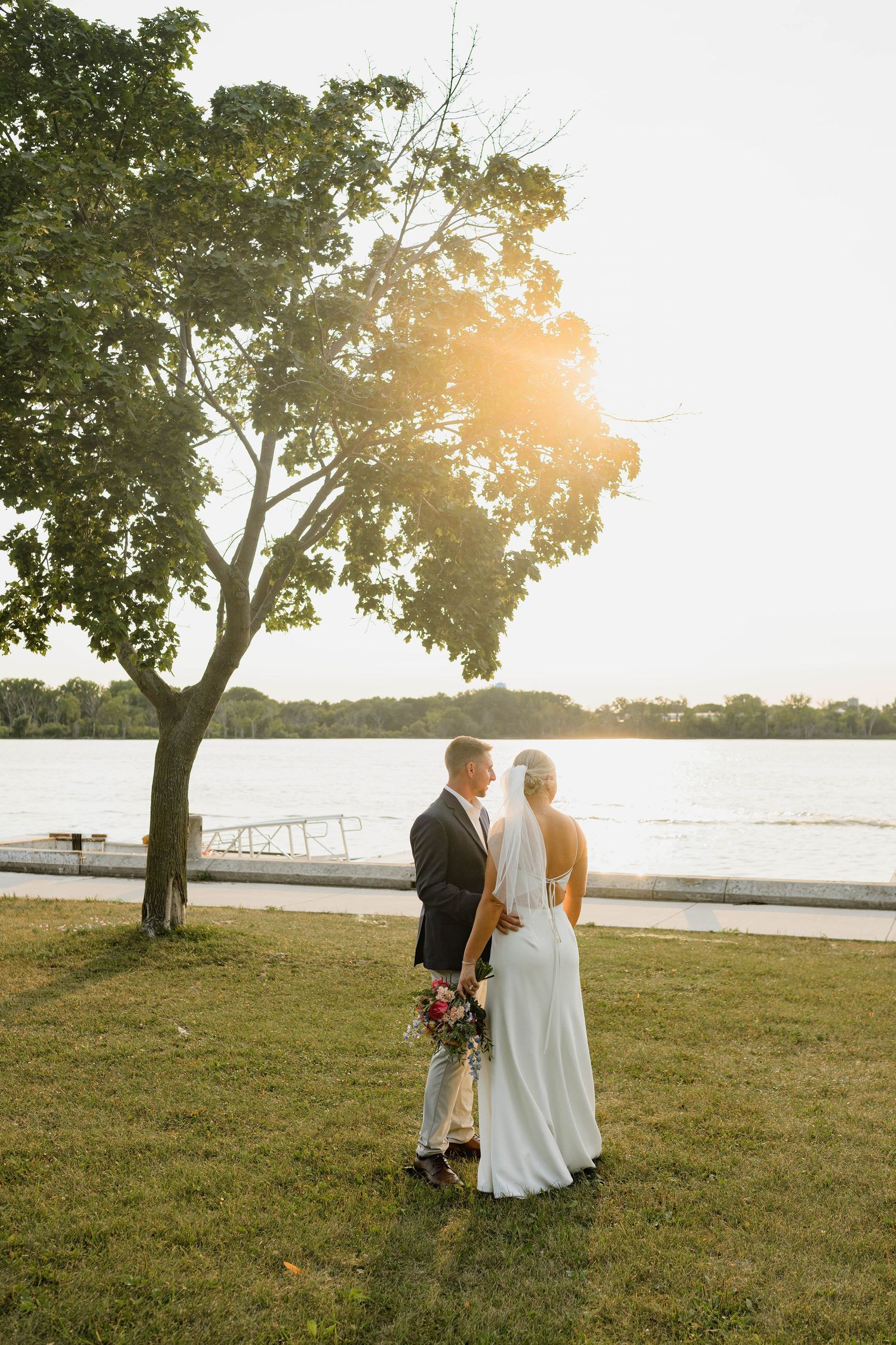 Bride and groom stand by a sunlit lakeside, bride holding a bouquet, surrounded by lush trees at golden hour.