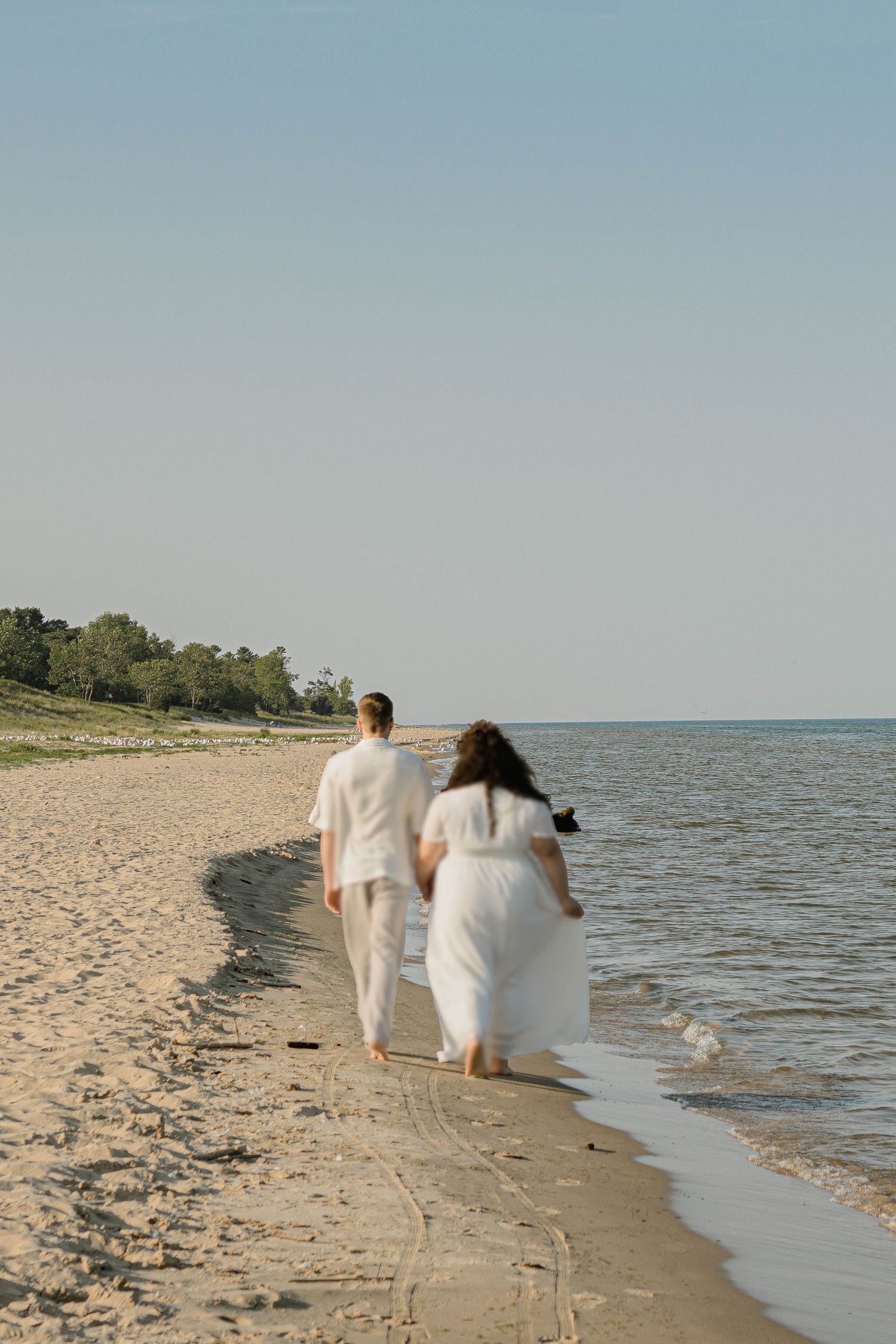 A couple in white walking along a sandy beach shoreline with calm water and trees in the background.