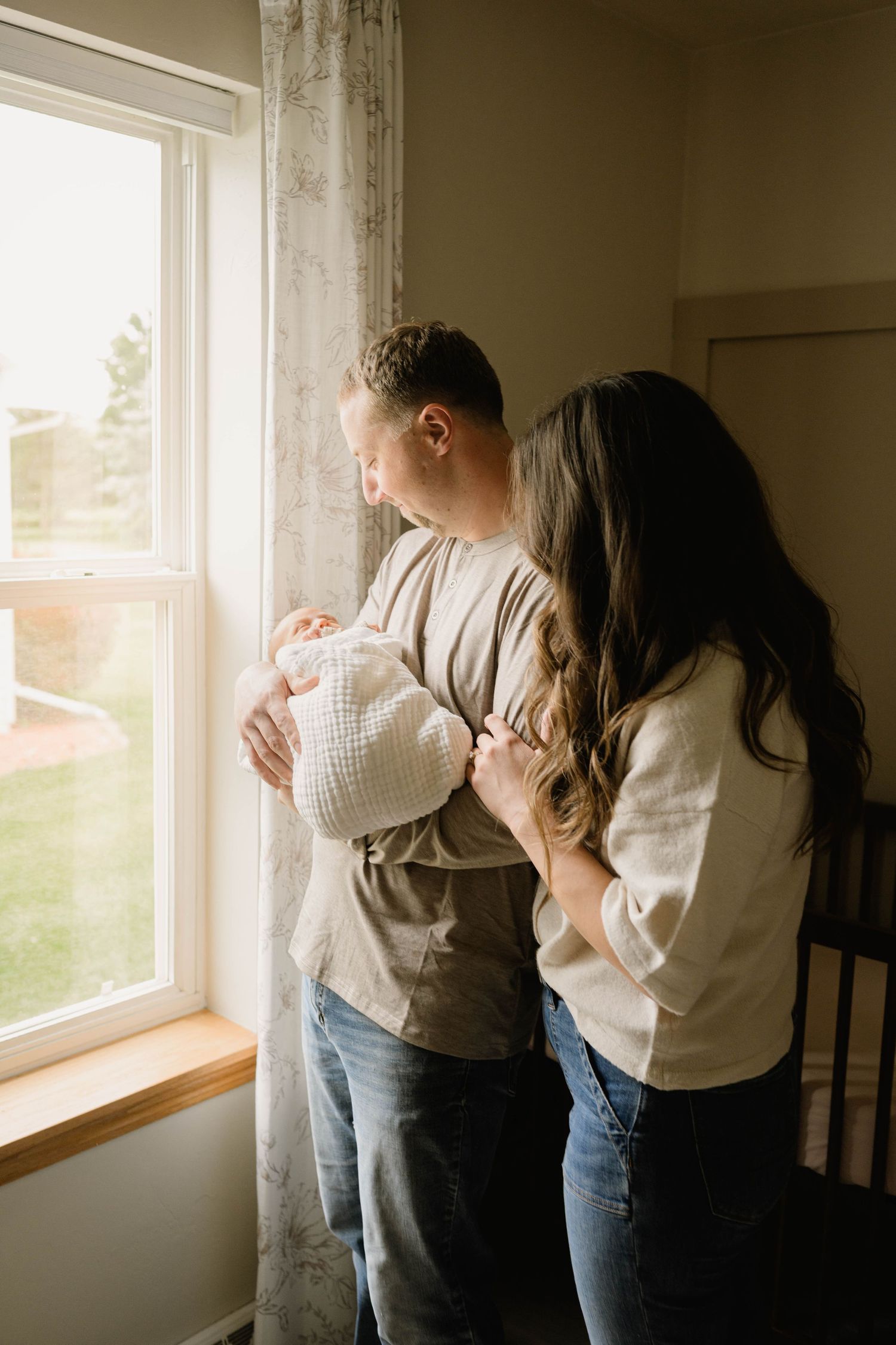 A couple stands by a sunlit window, the man cradling a newborn baby while the woman leans close in a warm home.