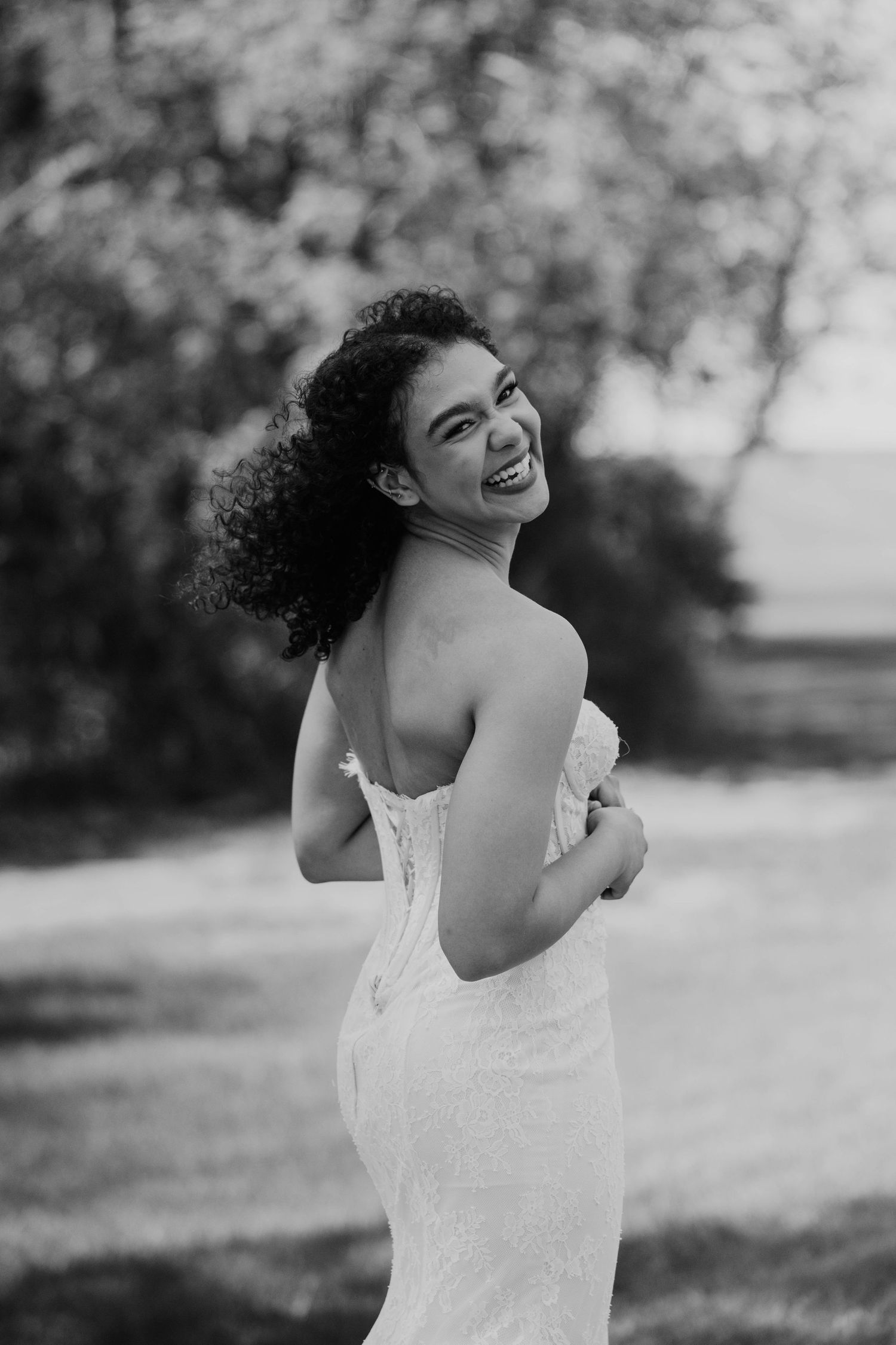 Black and white photo of a smiling bride in a strapless gown, glancing over her shoulder outdoors.
