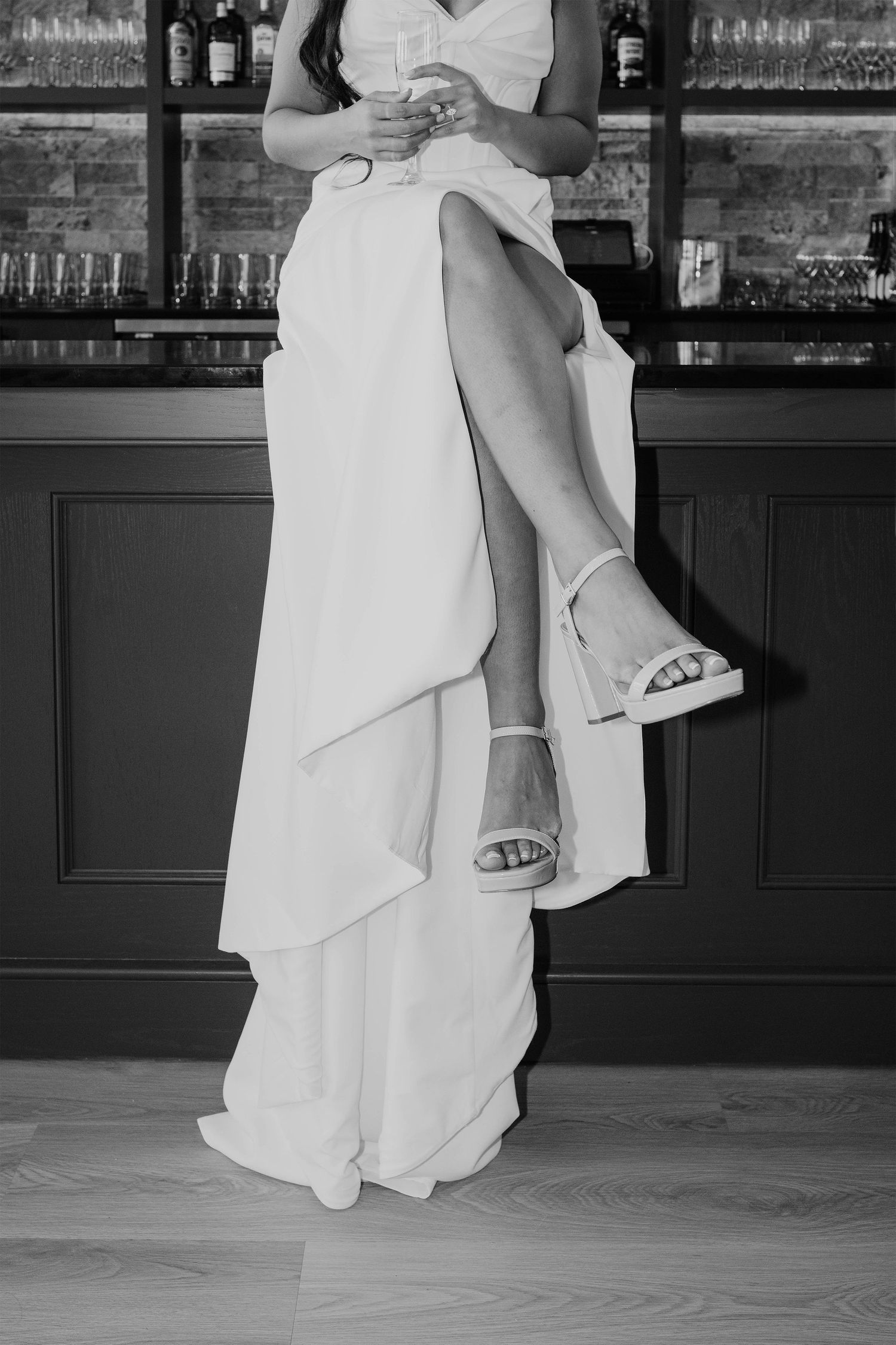 Black and white photo of a bride in a white gown with a thigh-high slit, wearing strappy heels, holding a glass at a bar.