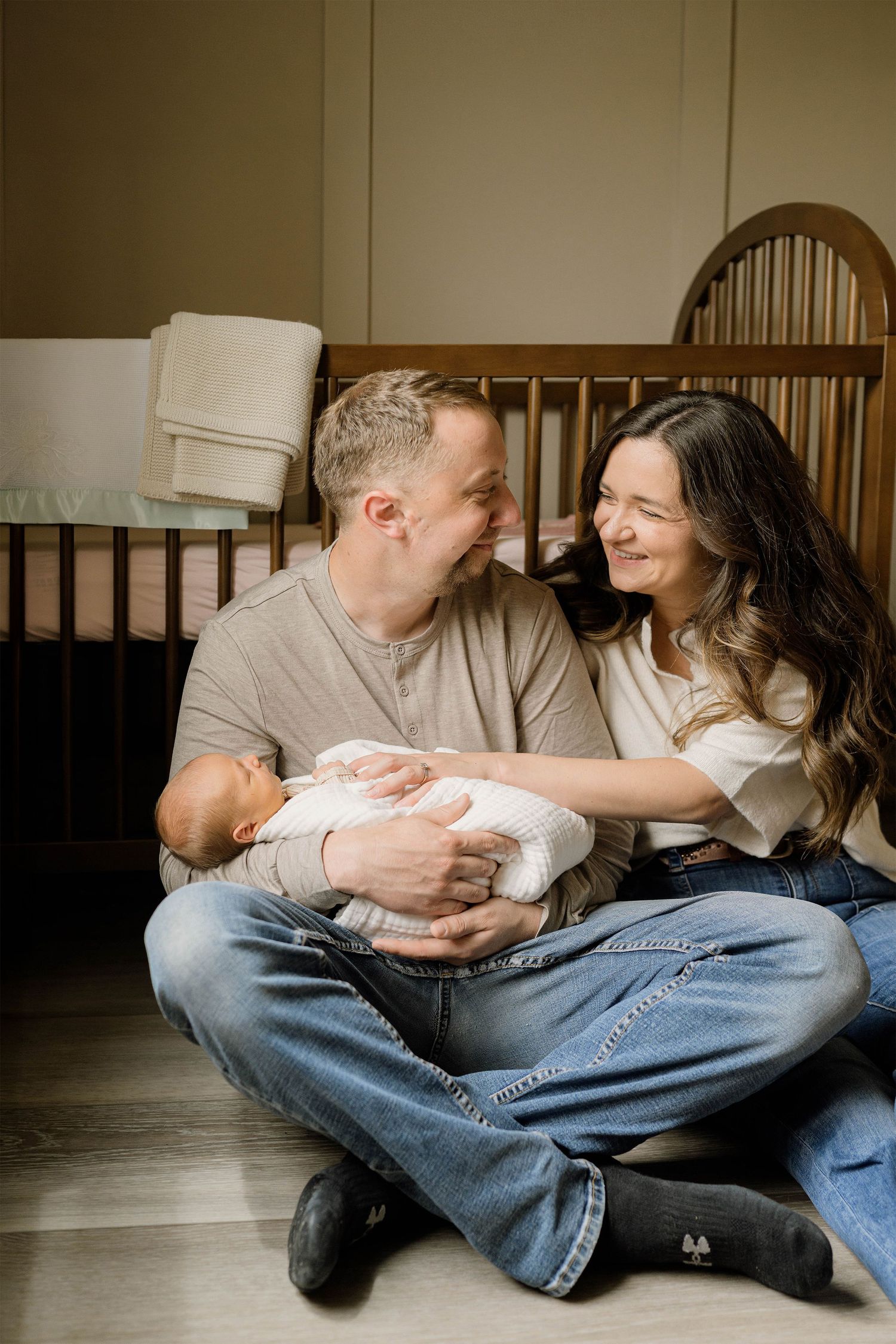 Happy couple sitting on nursery floor with newborn baby, smiling at each other beside a wooden crib.
