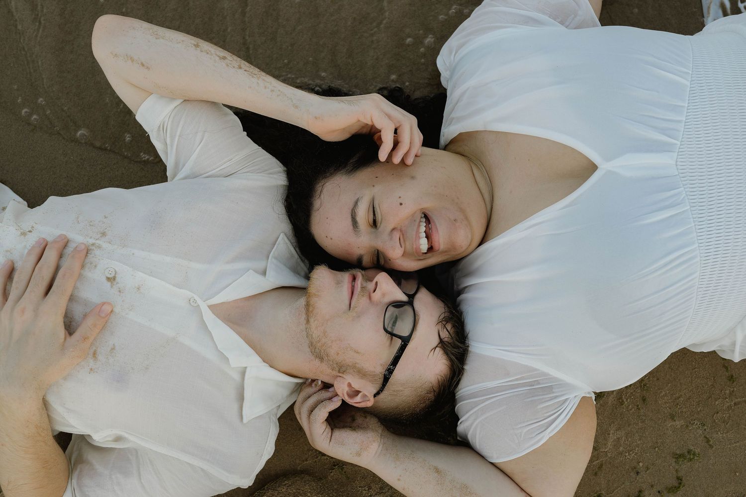 A smiling couple lies on the sand looking at each other, both dressed in white outfits.
