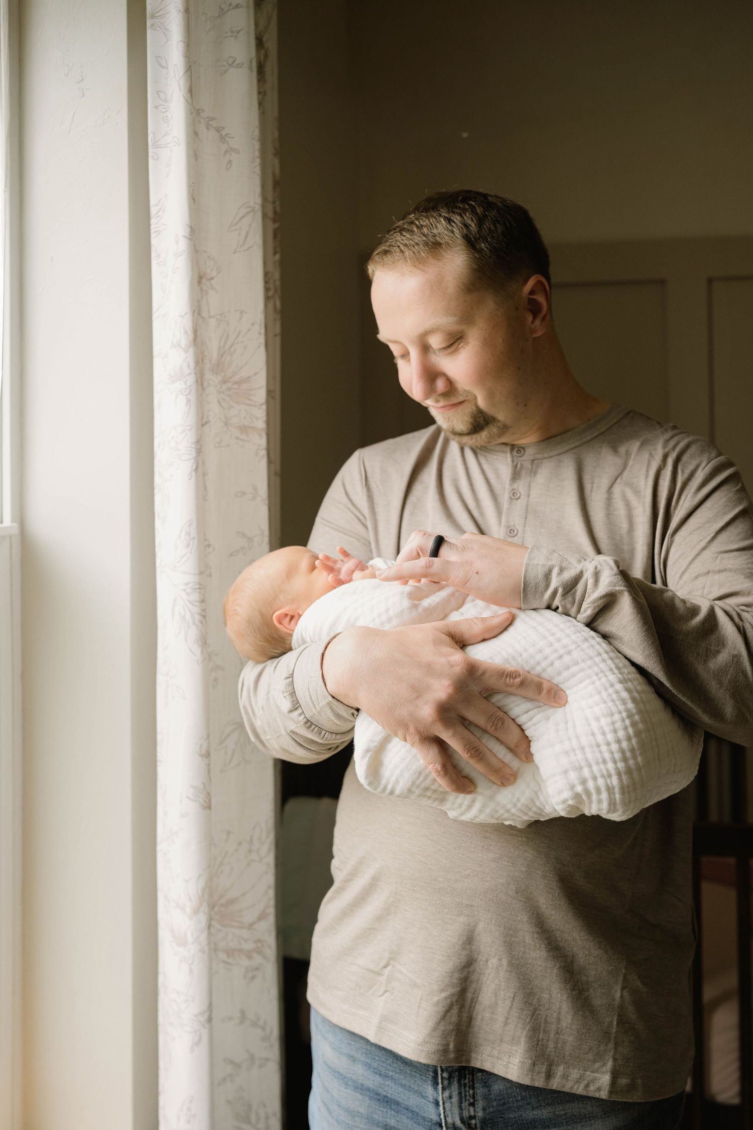 New father stands by a window gazing lovingly at his swaddled newborn baby in his arms.