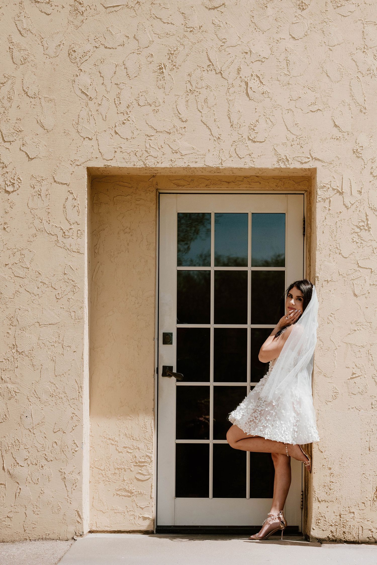 Bride in short white lace dress and veil posing by a French door against a textured beige stucco wall.