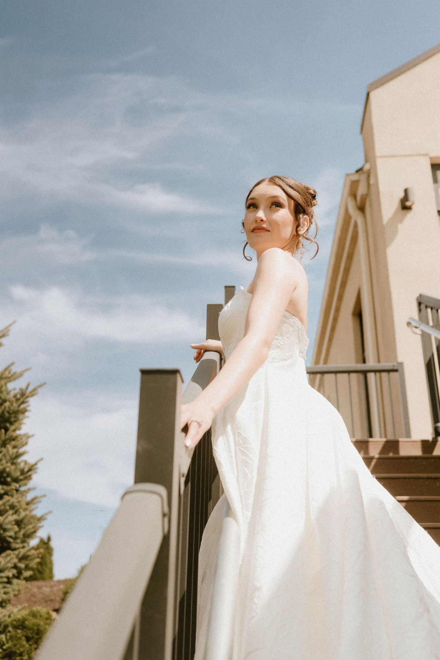 Bride in white gown standing on outdoor staircase, flowing dress caught in breeze against blue sky.