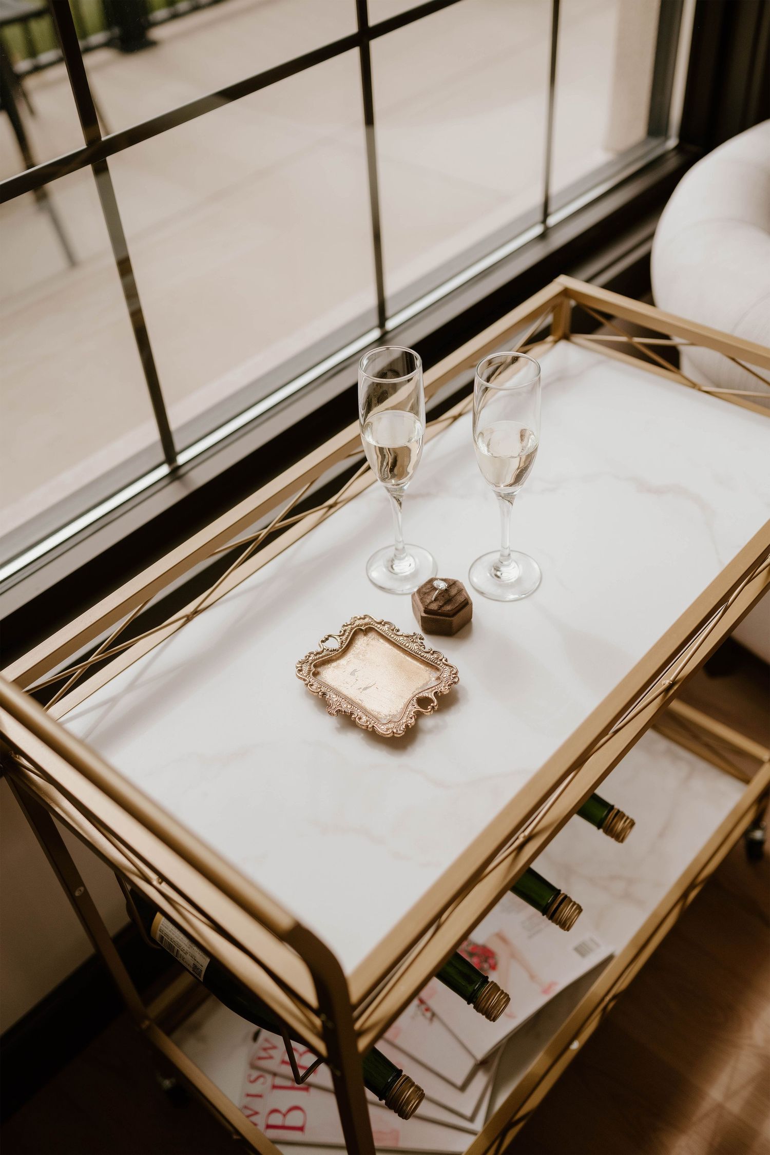 Gold bar cart with marble top holding two champagne glasses, chocolates, and a ring box near a large window.