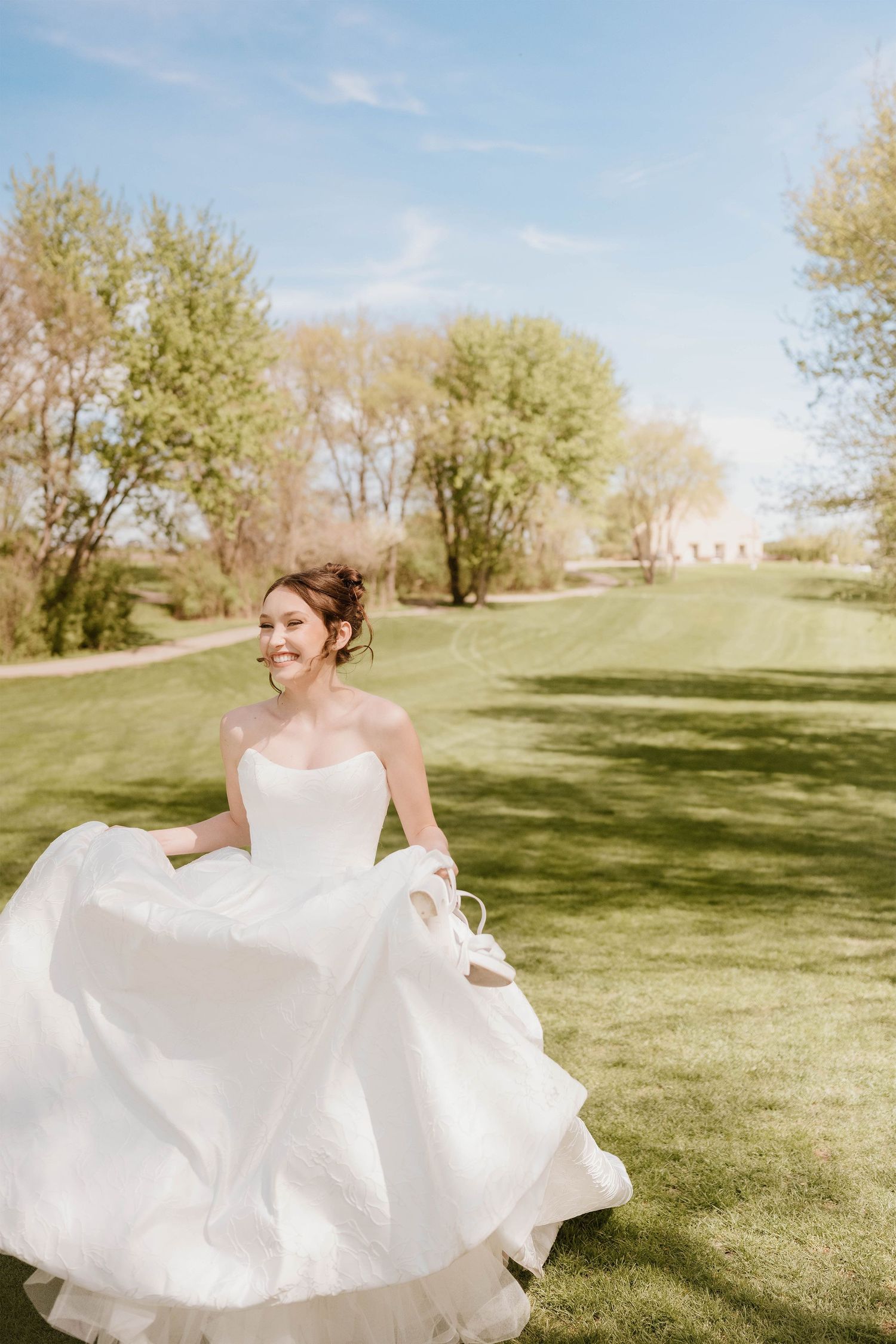 Smiling bride in a flowing white strapless ball gown twirling on a lush green lawn with trees and blue sky.