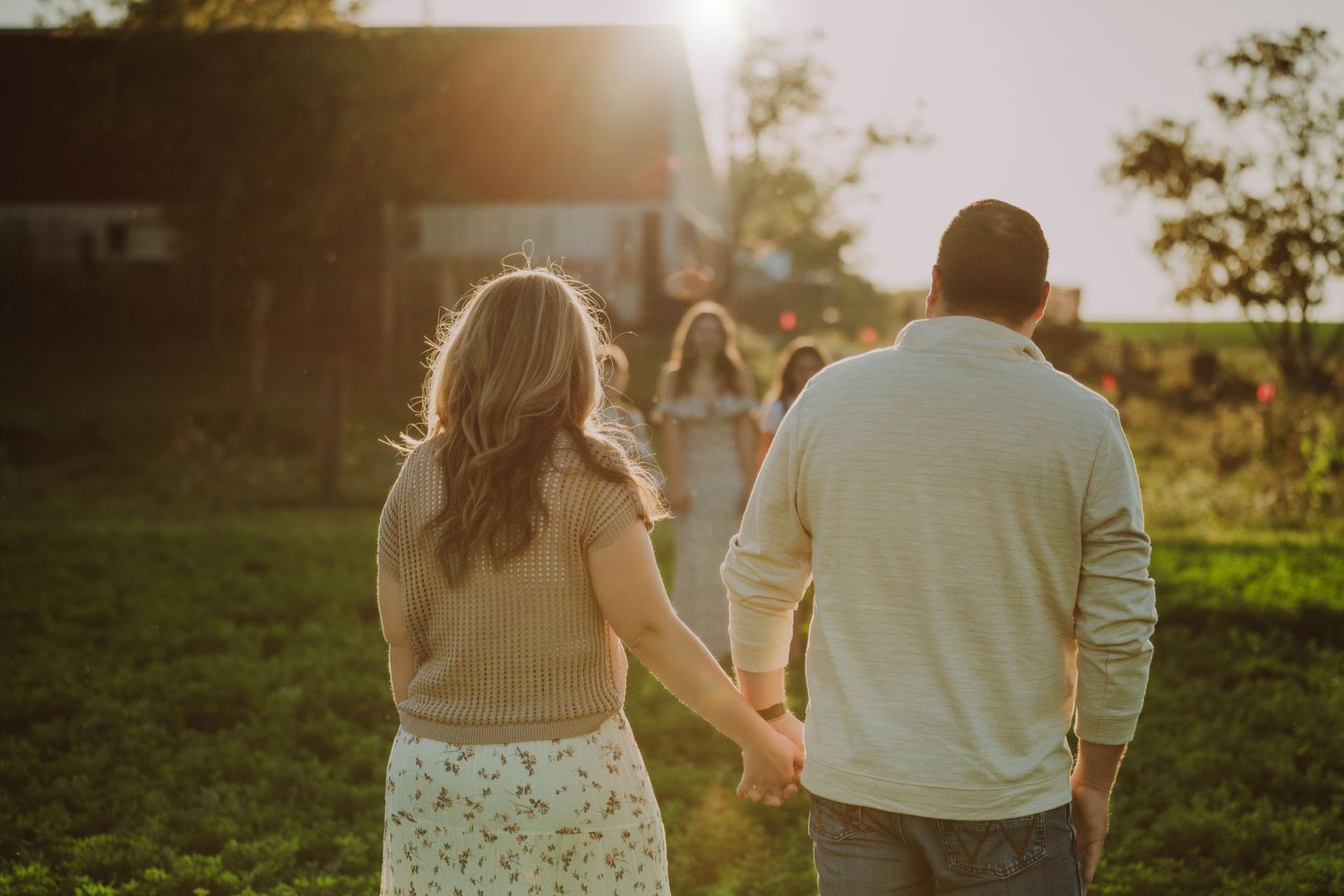 A couple holds hands walking through a golden sunlit field at sunset creating a romantic silhouette.