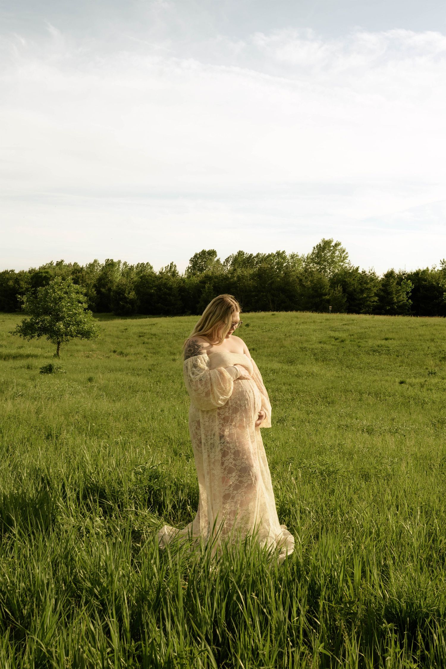 Pregnant woman in white lace gown standing in lush green field with trees in background.