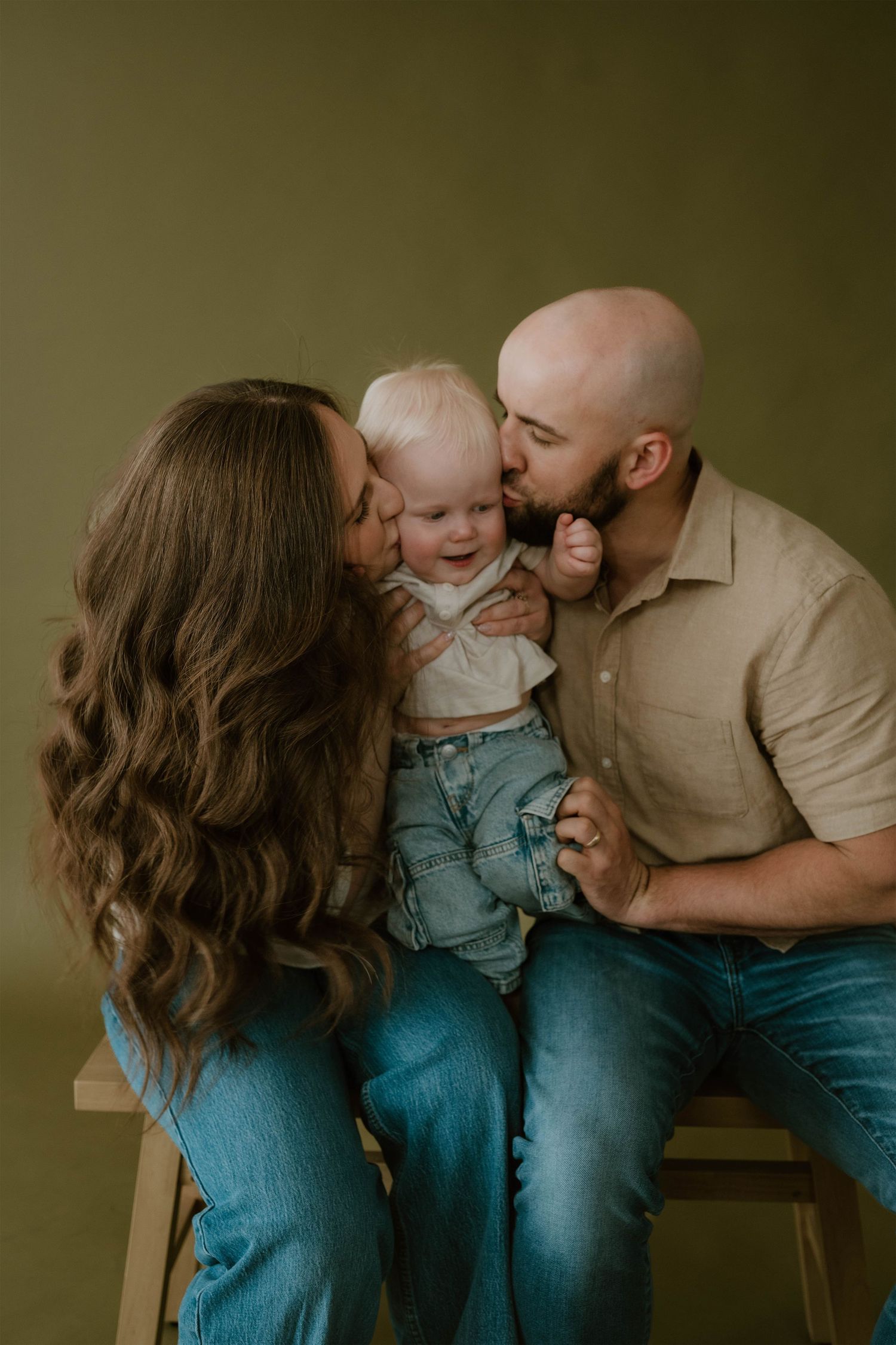 A loving family portrait with parents kissing their smiling baby on a warm olive green studio background.
