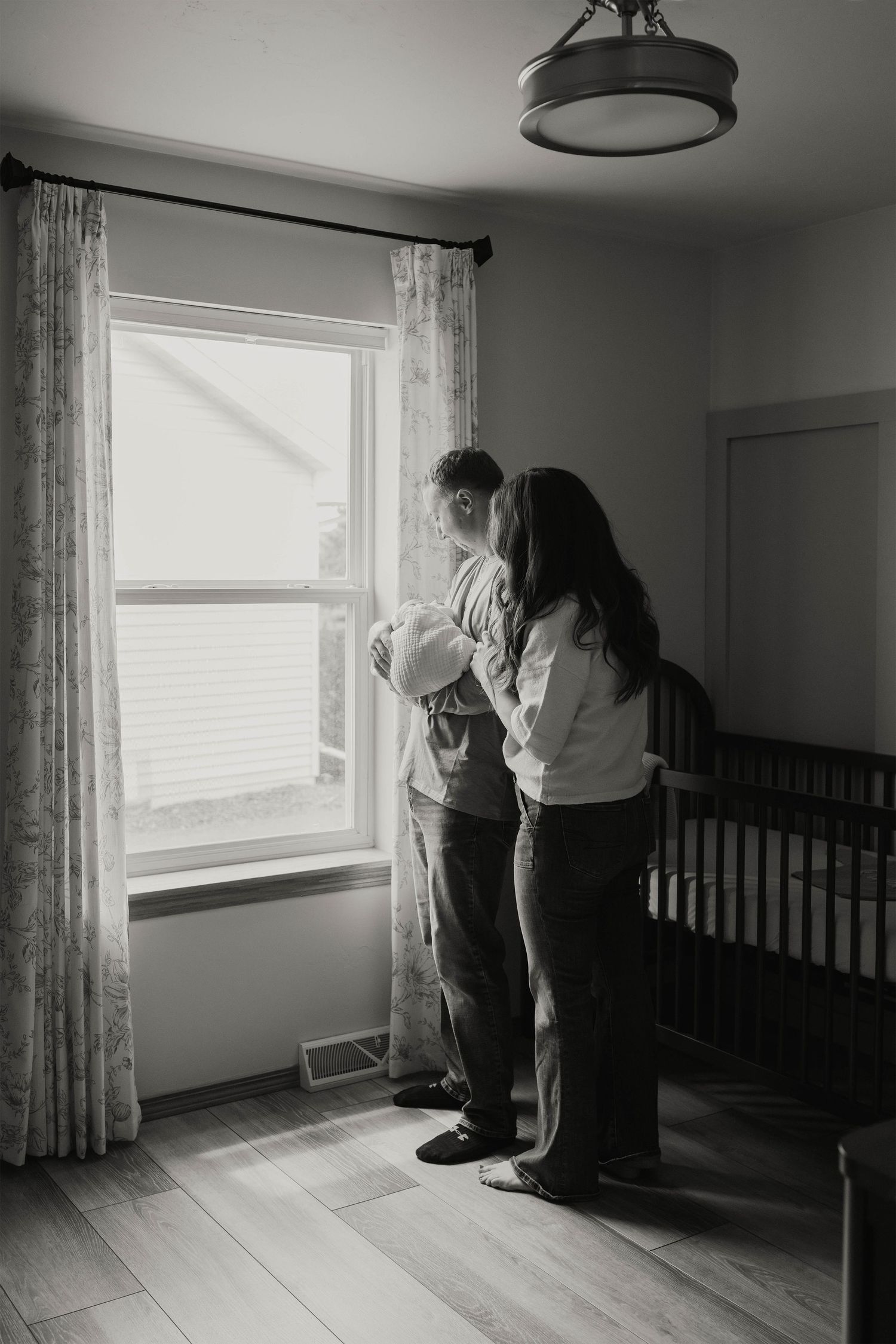 Black and white photo of parents holding newborn baby by nursery window with crib nearby.