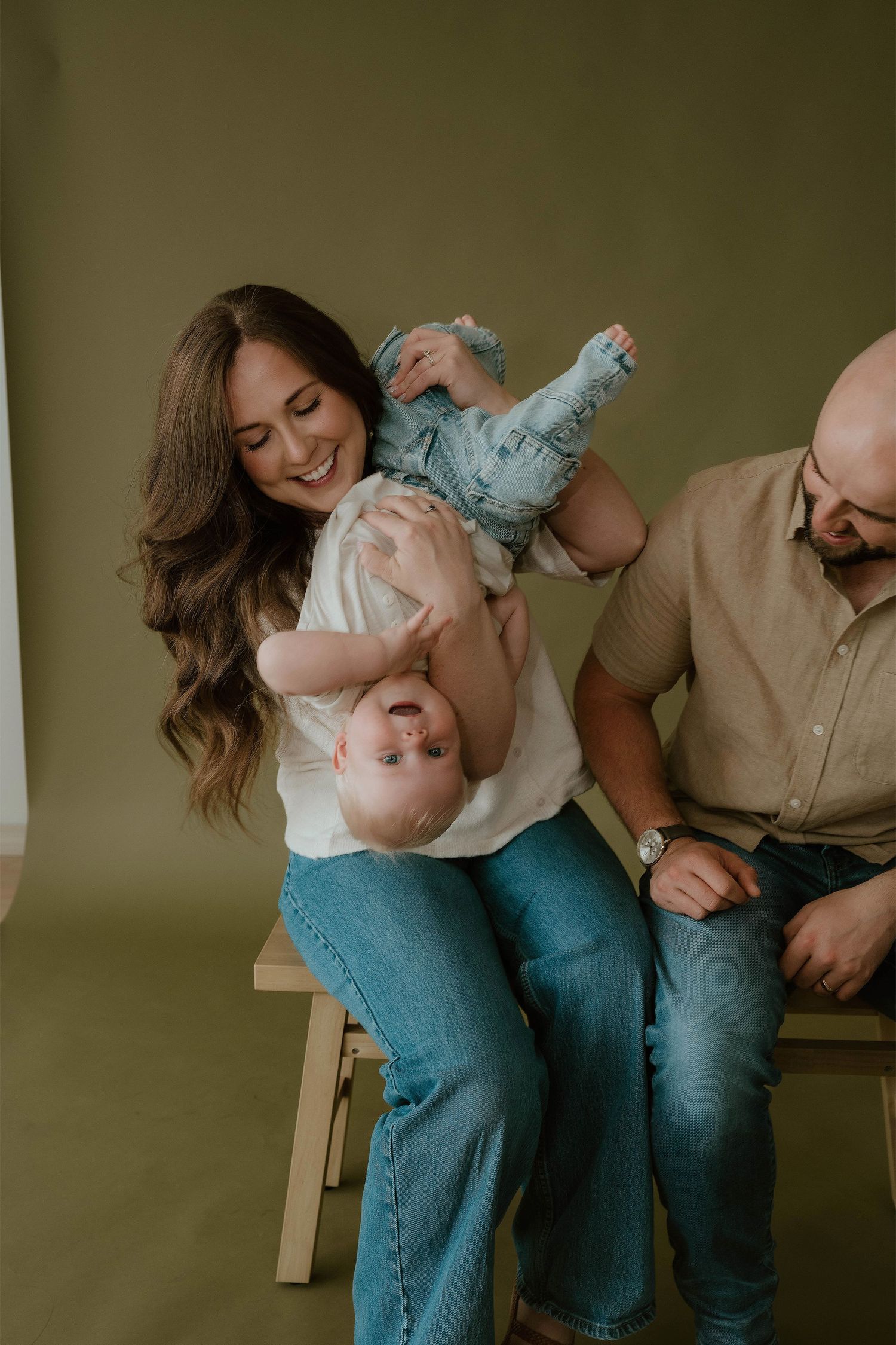 A smiling mother holds her baby upside down while a father looks on, all seated against an olive green studio backdrop.