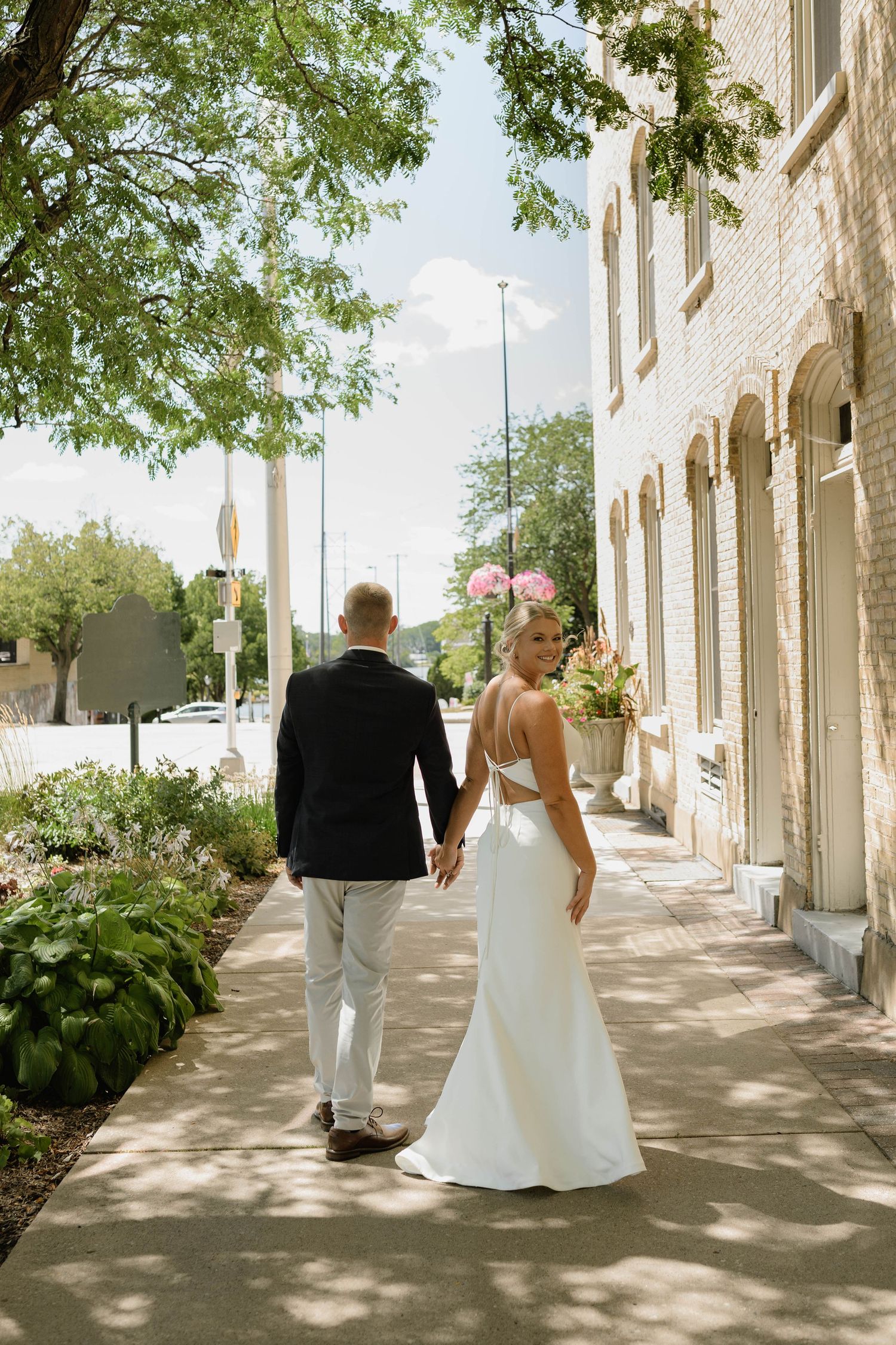 A bride and groom walk hand in hand down a sunny sidewalk, the bride wearing a backless white gown with a floral hair accessory.