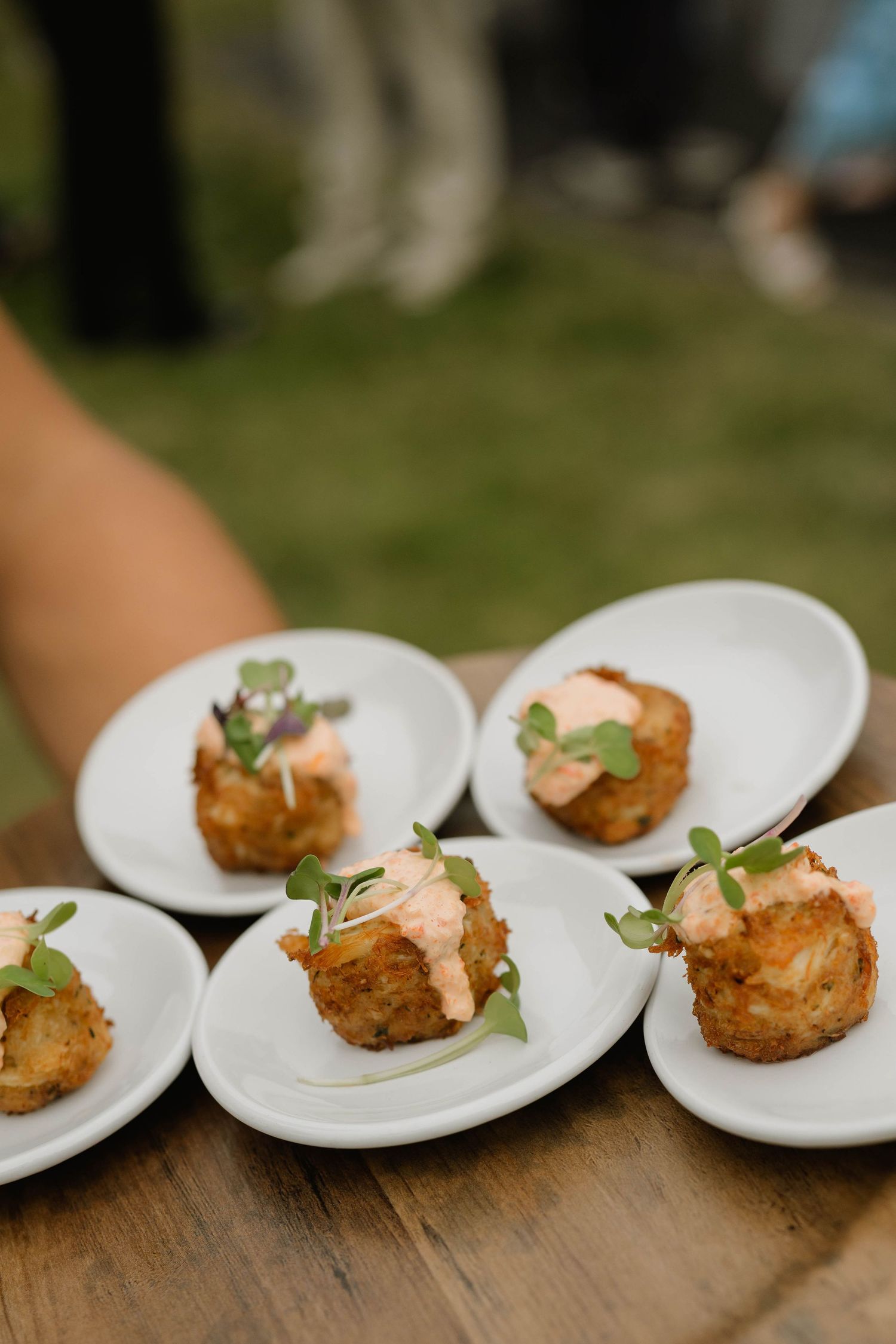Elegant appetizers on white plates featuring crispy crab cakes topped with pink sauce and fresh microgreens at an outdoor event.