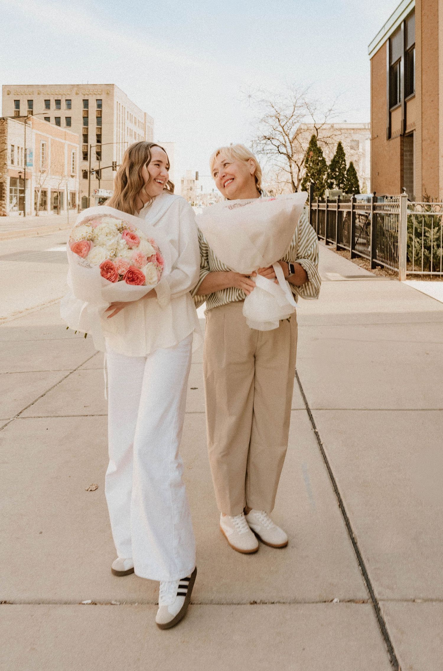 Two smiling women standing outdoors holding large wrapped floral bouquets in a sunny urban setting.