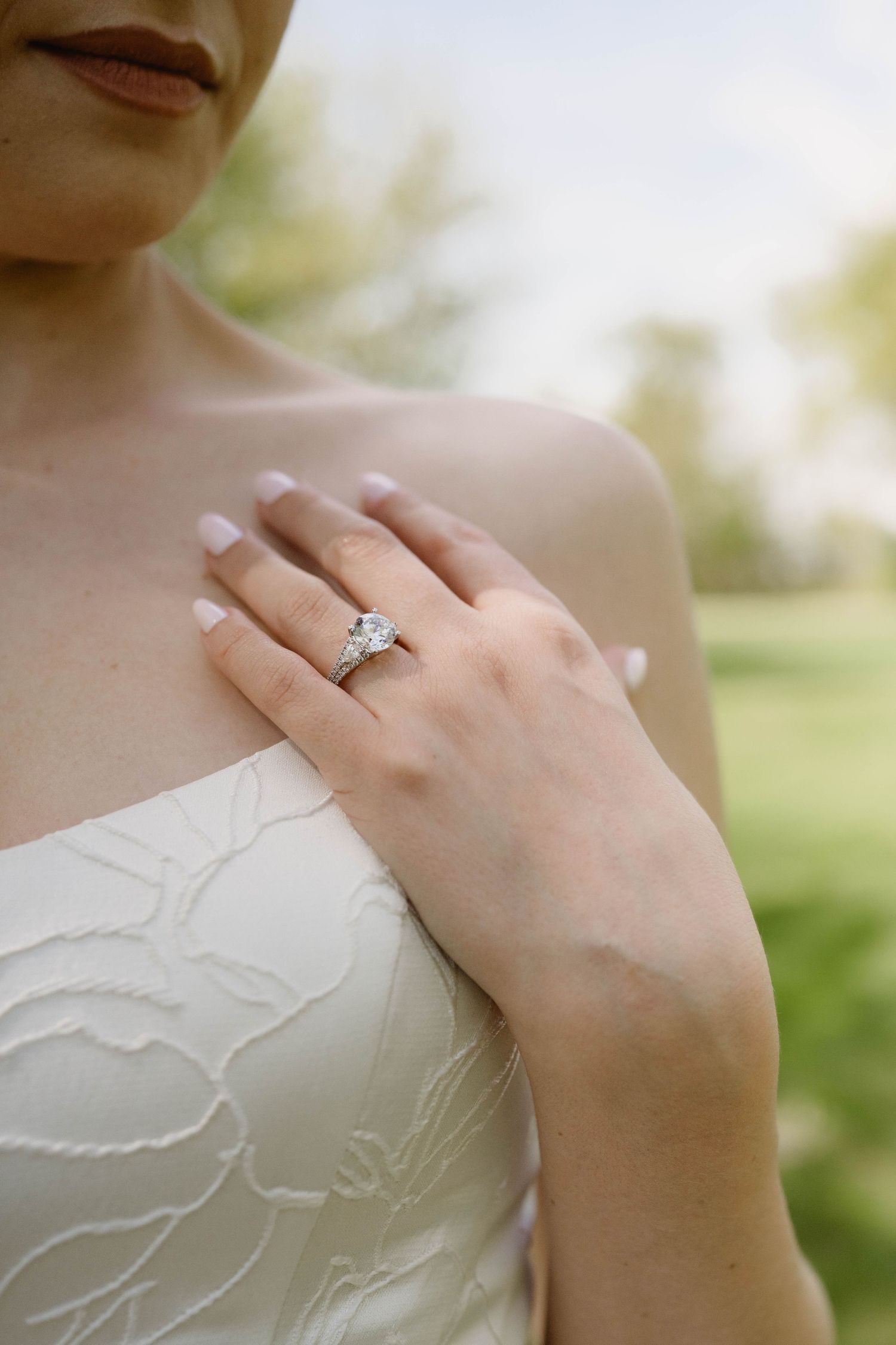 Bride in white lace gown showcasing a sparkling diamond engagement ring on her hand placed over her chest.