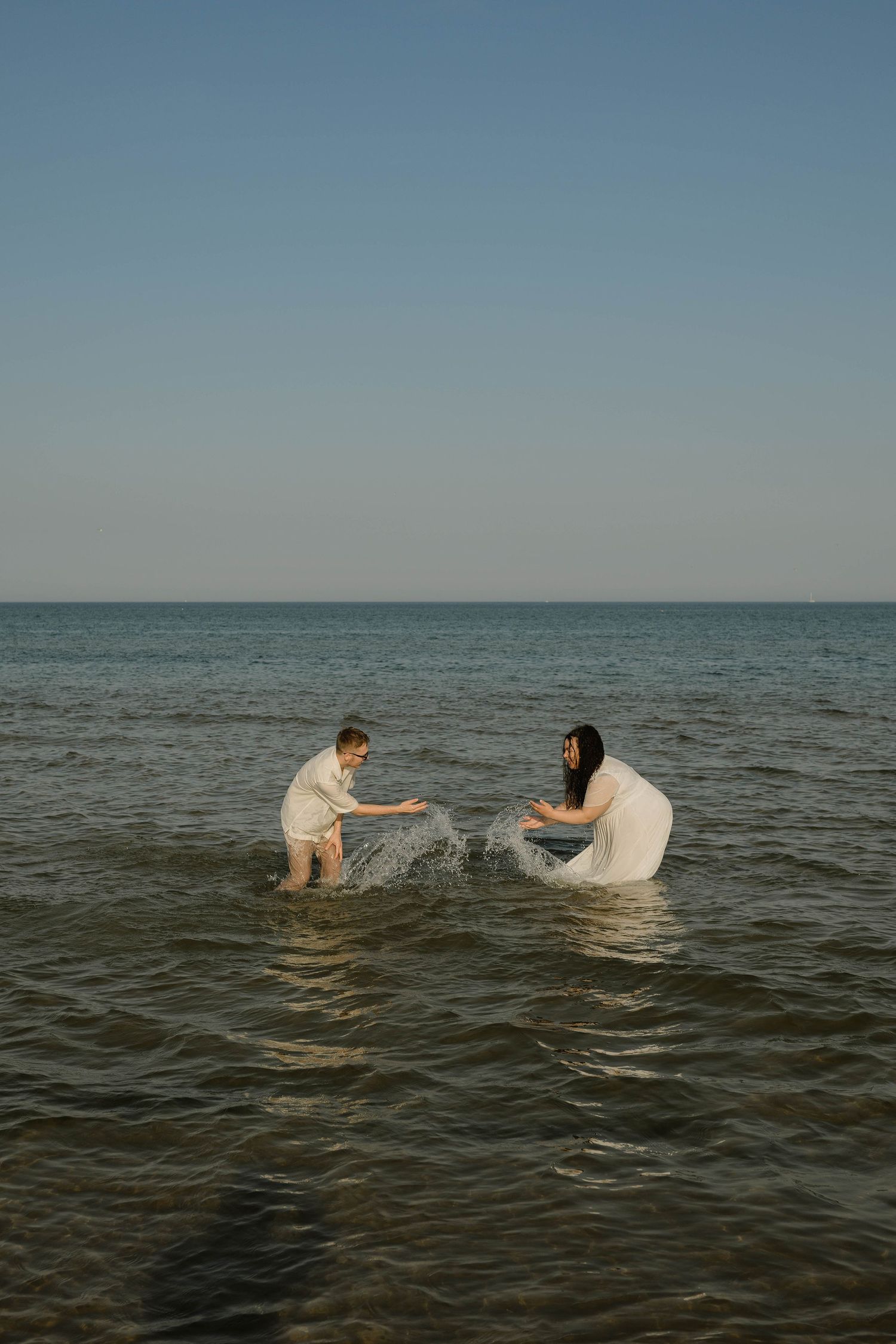 Couple splashing water at each other while standing in the ocean wearing white clothes.