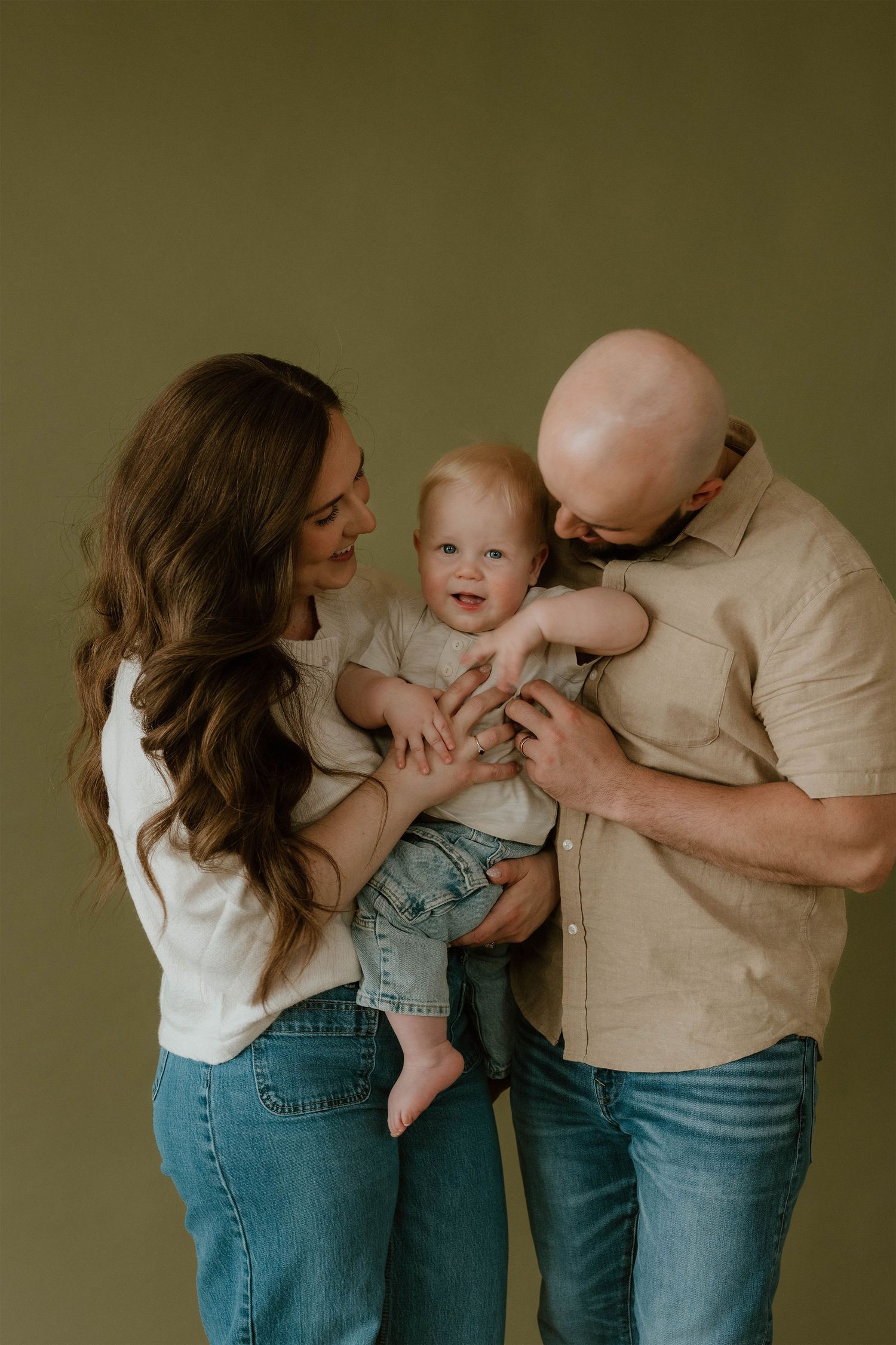 Family portrait of parents holding their smiling baby in a studio with olive green background.