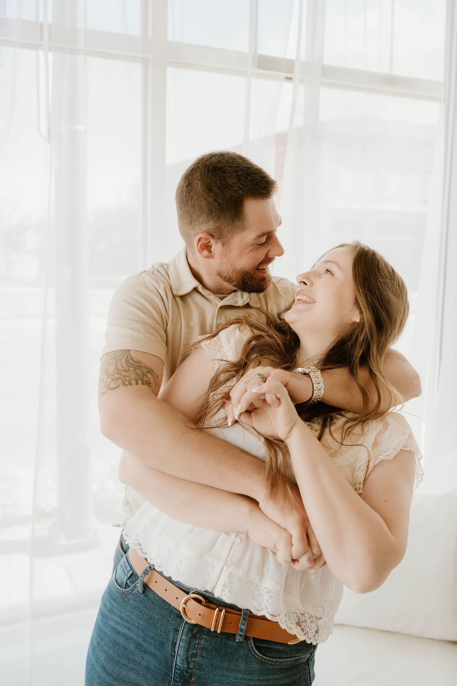 A happy couple embraces and smiles at each other in a bright, airy studio with white curtains.