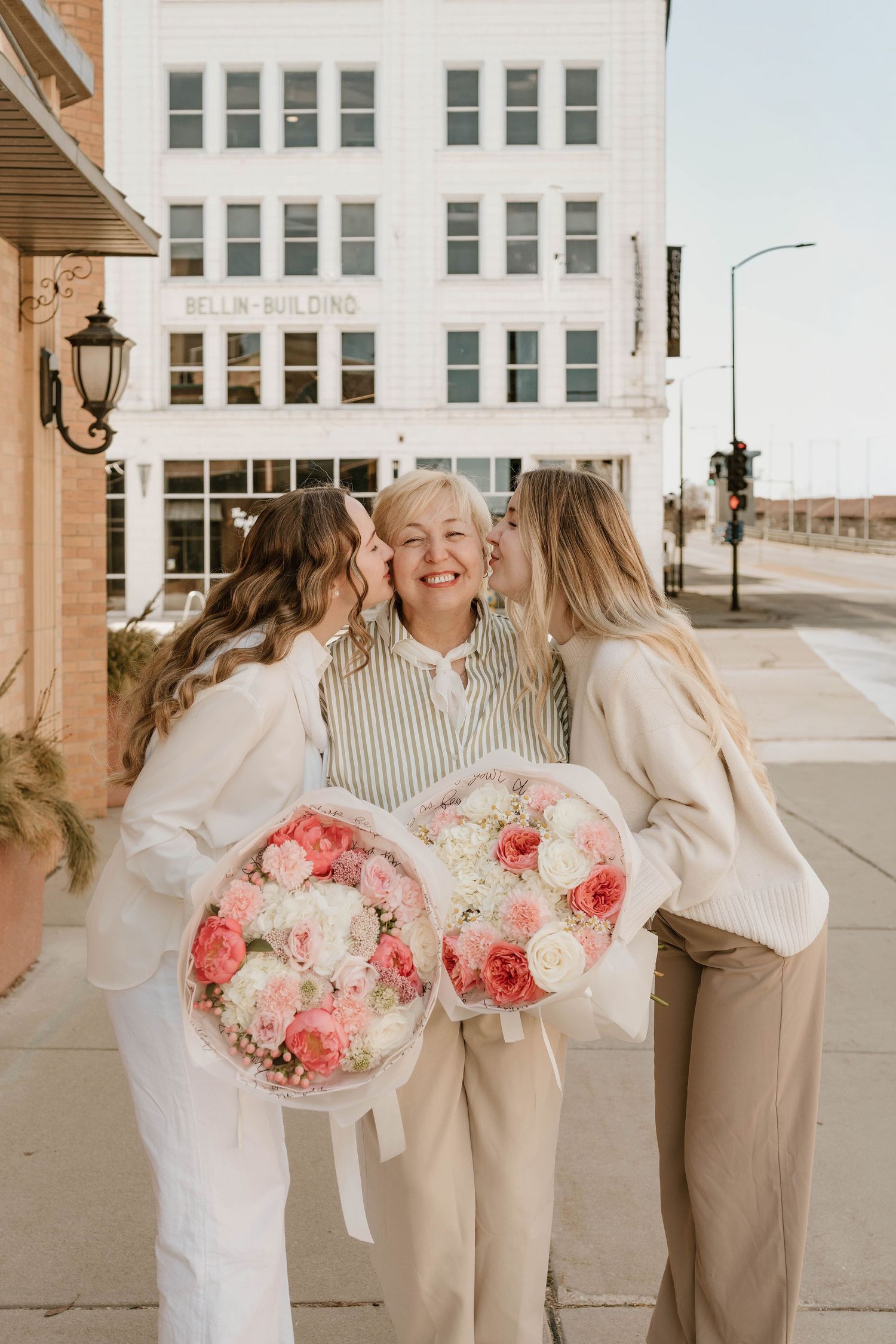 Two daughters kissing their smiling mother on the cheeks while holding pink and white flower bouquets outdoors.