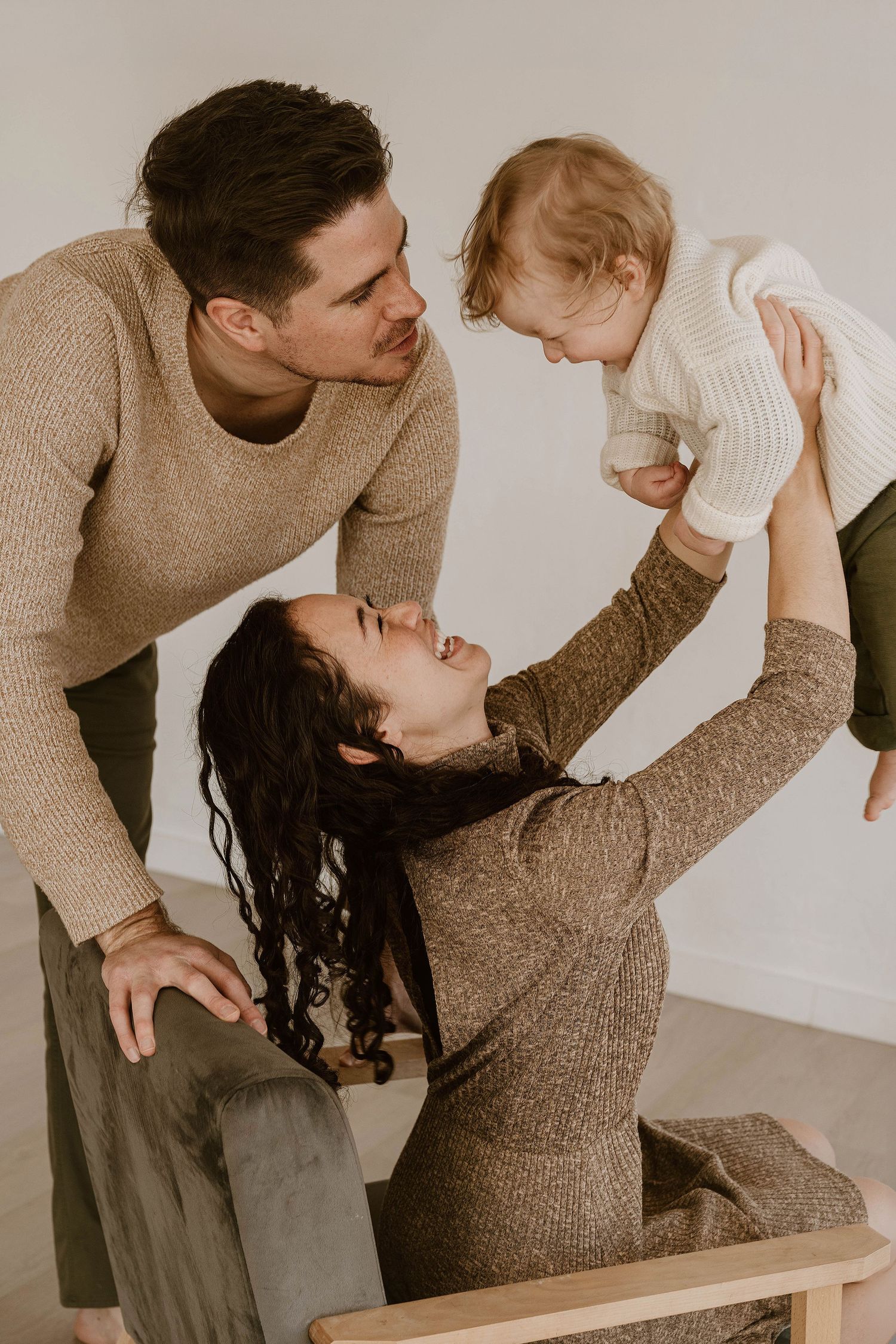 A family shares a tender moment together wearing cozy neutral sweaters against a white background.