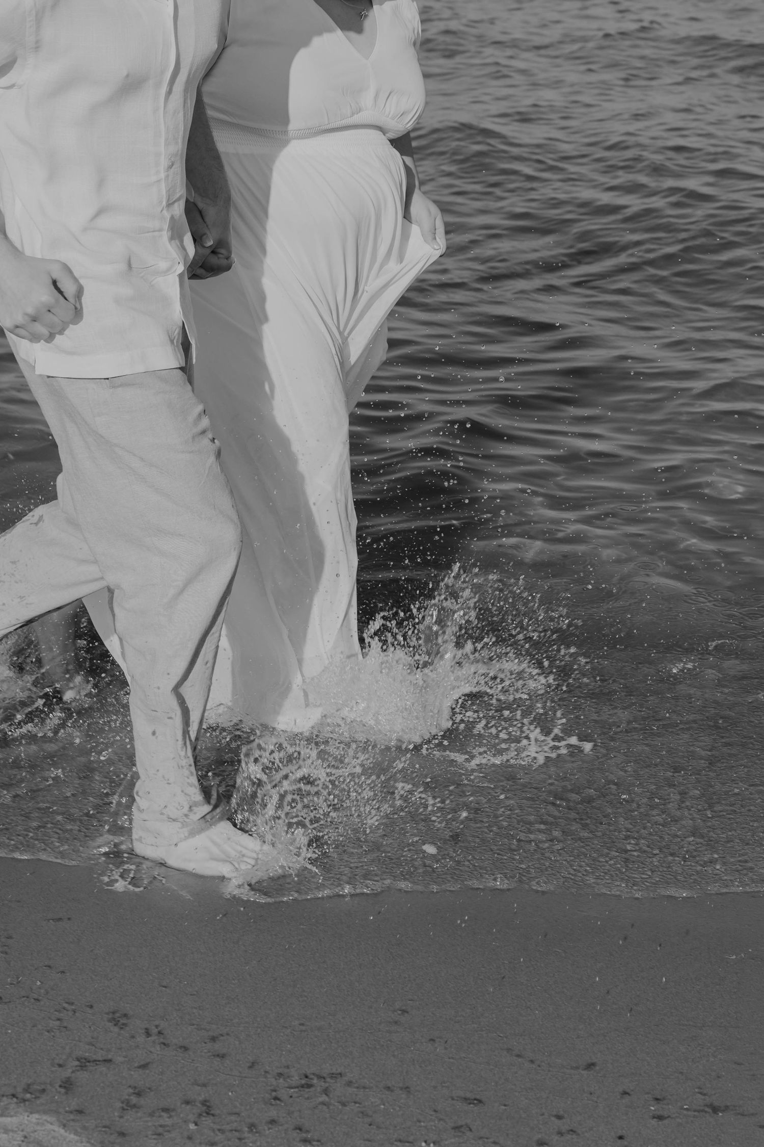 Black and white photo of couple running through ocean waves on a sandy beach.