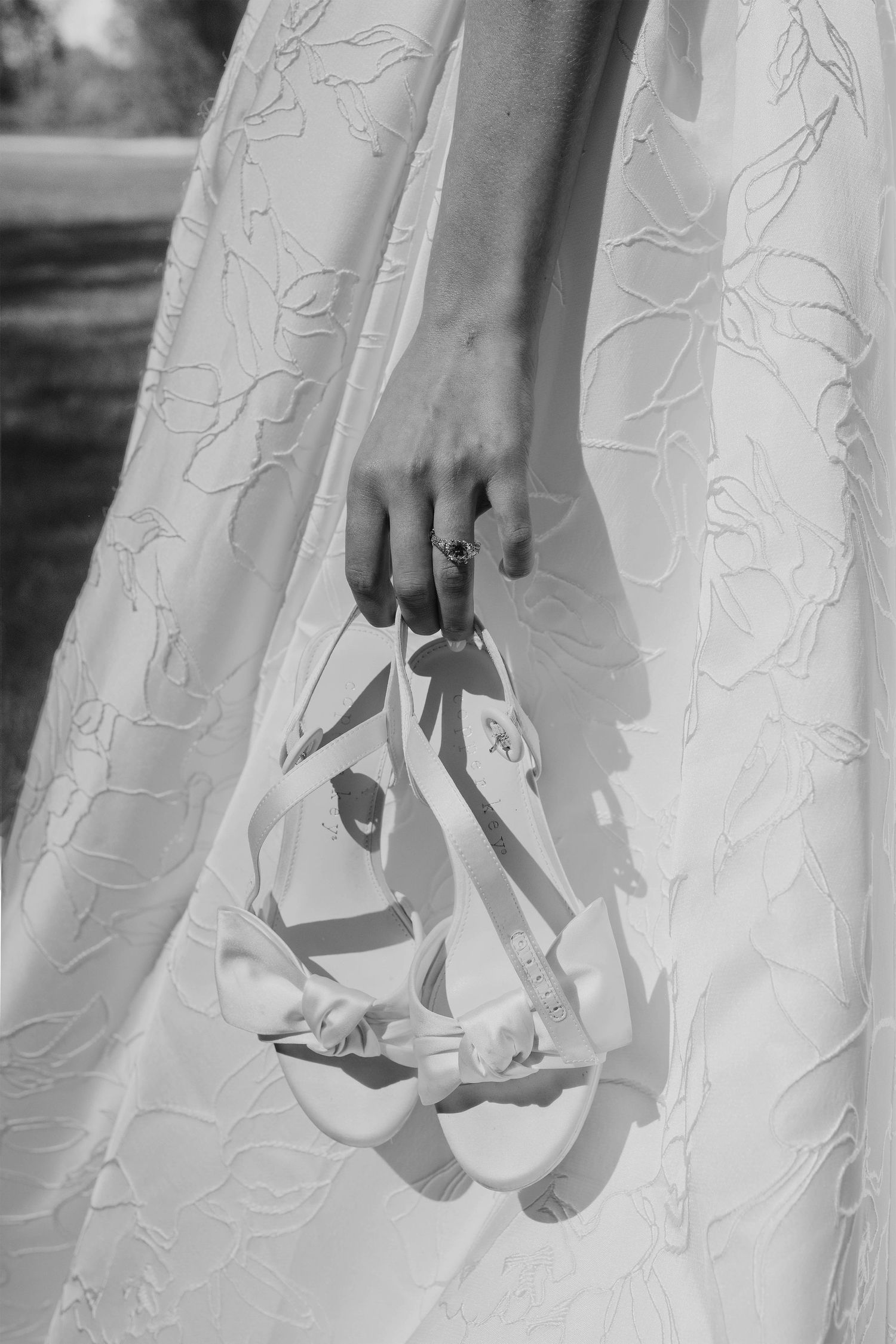 Bride in floral lace gown holding white satin sandals, black and white wedding photography.