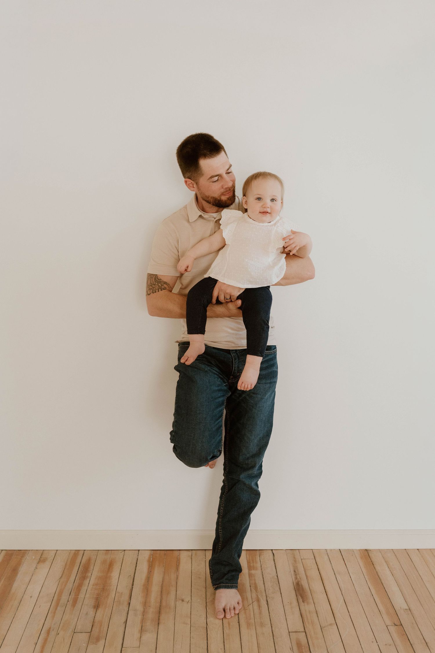A tattooed father kisses his baby while standing against a white wall on hardwood floors.