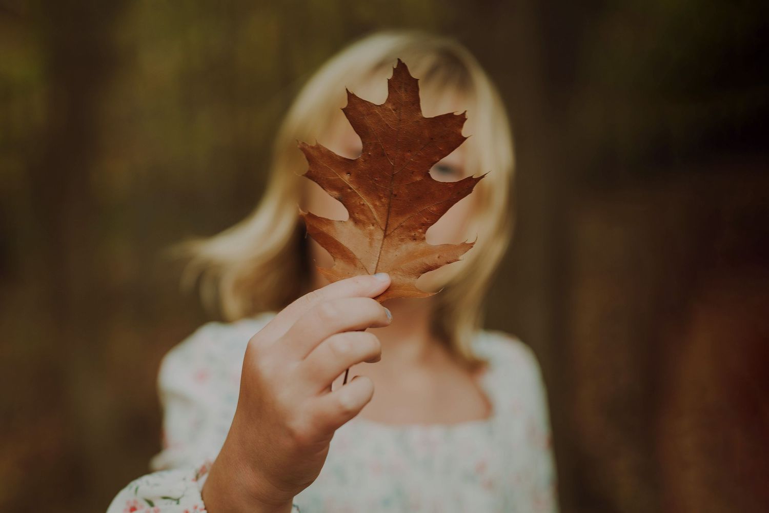 Person holding autumn maple leaf in front of their face against blurred outdoor background.