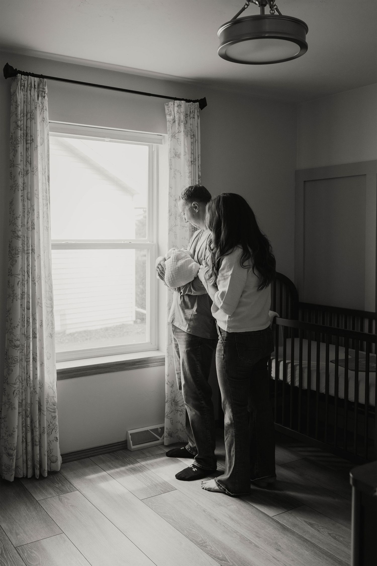 Black and white photo of new parents holding newborn baby by nursery window with crib nearby.