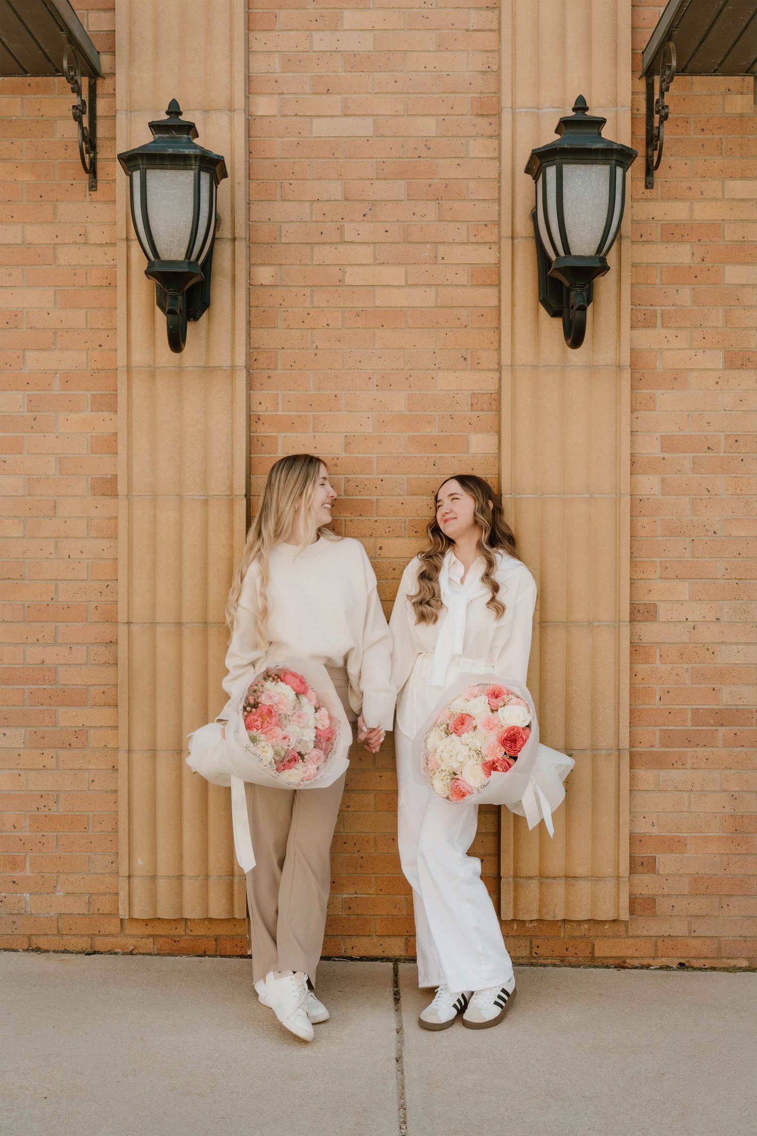 Two brides in white outfits holding pink floral bouquets, smiling at each other outside a brick building.
