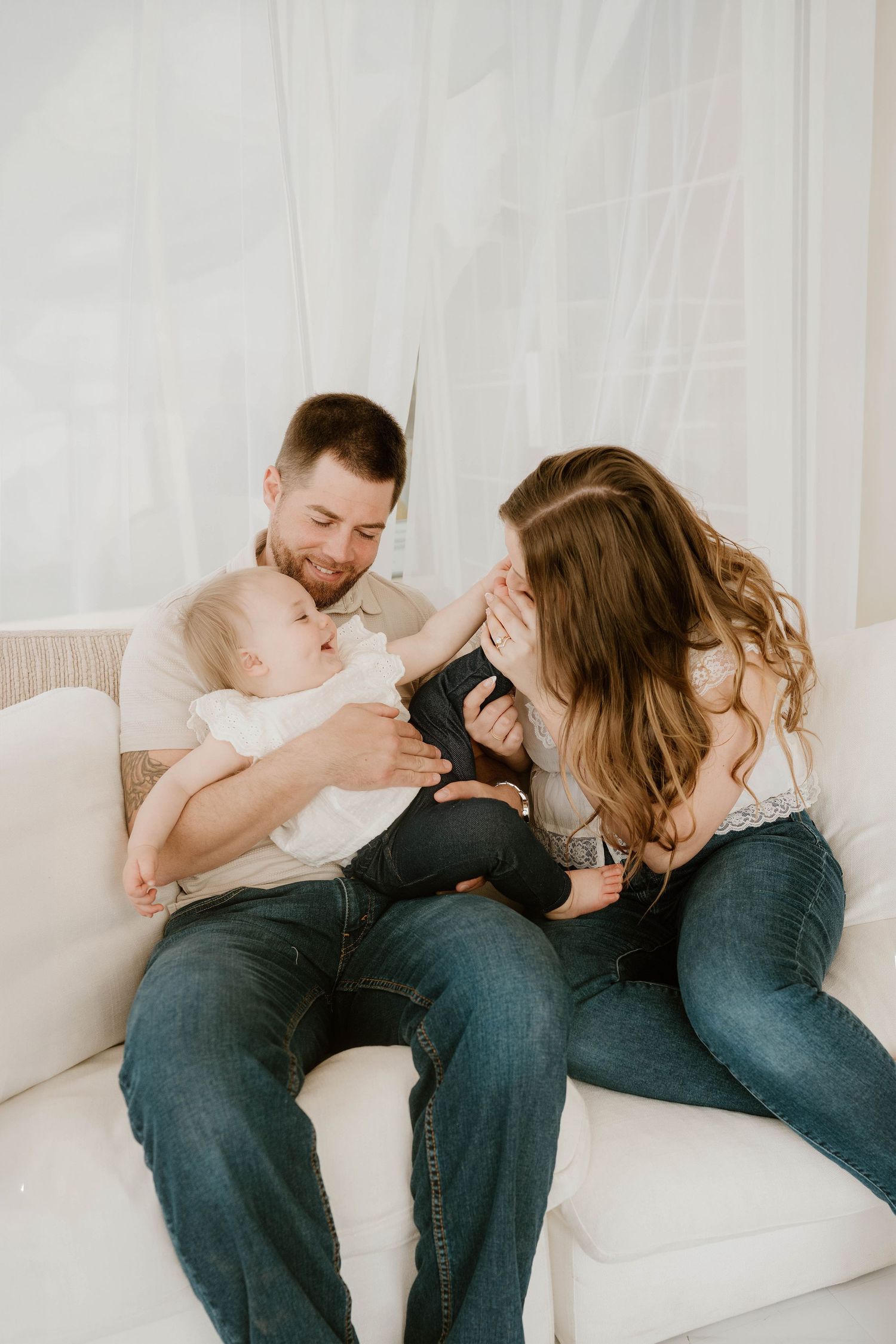 A happy young family with a baby sits on a white couch, sharing a joyful moment together.