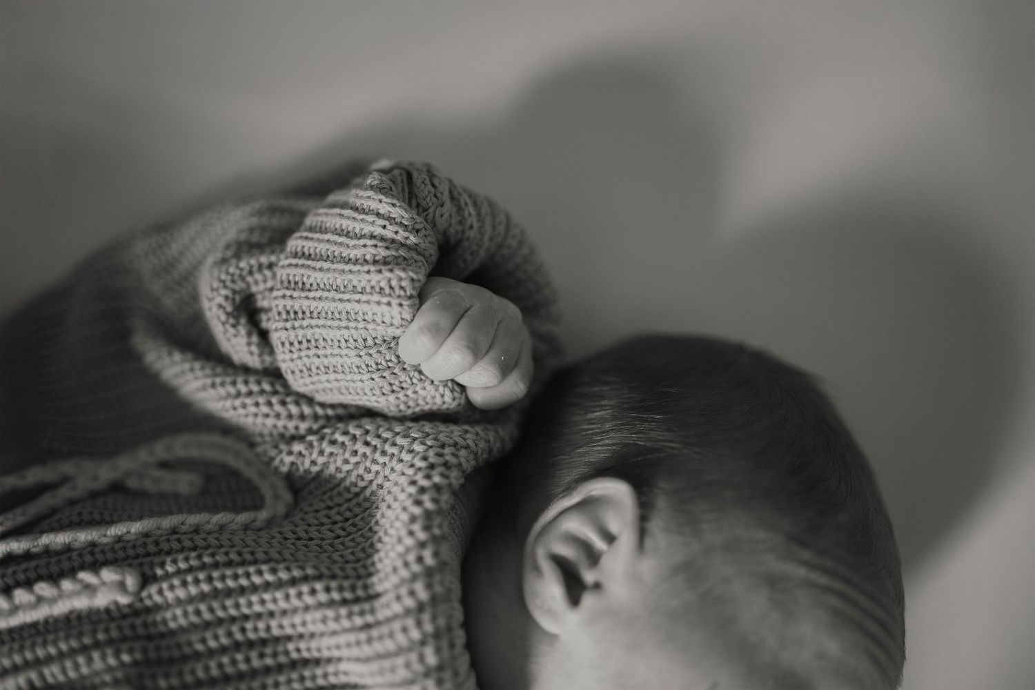 Black and white photo of a sleeping newborn baby wrapped in a knit blanket, tiny hand visible near face.